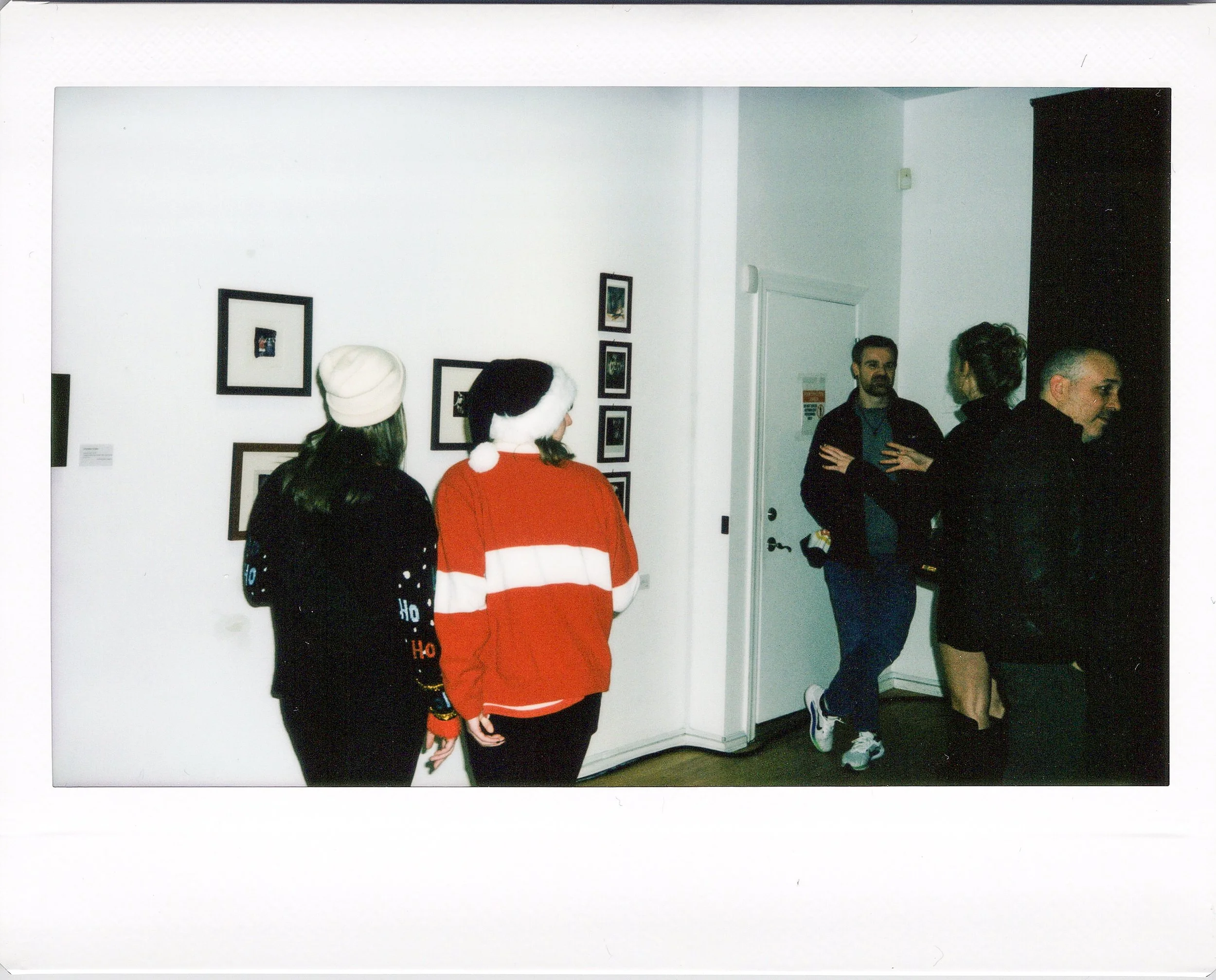 People viewing artwork in a Chicago pop-up gallery exhibition, with some wearing holiday-themed clothing, such as Santa hats and Christmas sweaters.
