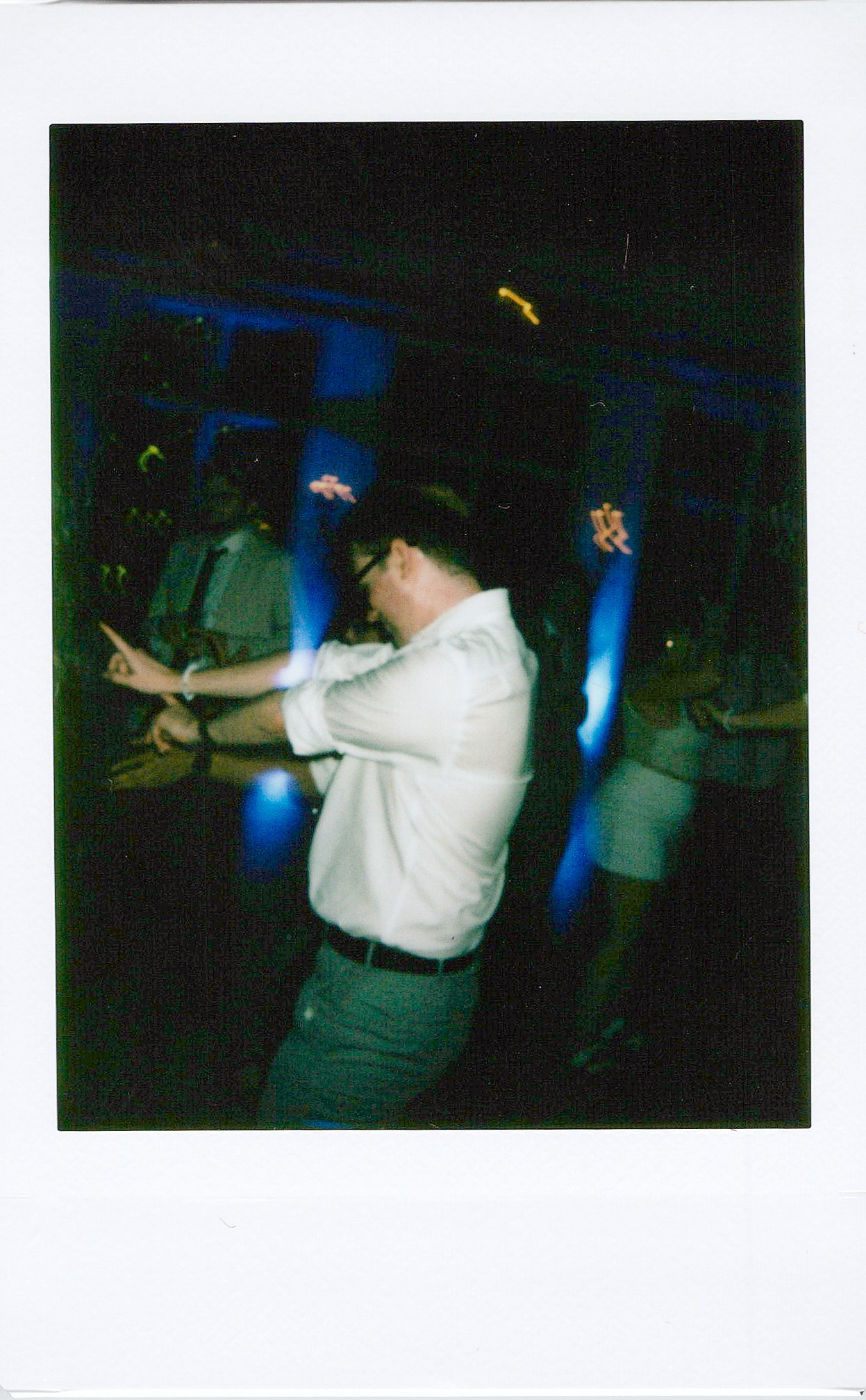 A man with glasses wearing a white shirt and green pants is standing with his head bent down and arms crossed in a dark room with blue and yellow dotted lights.