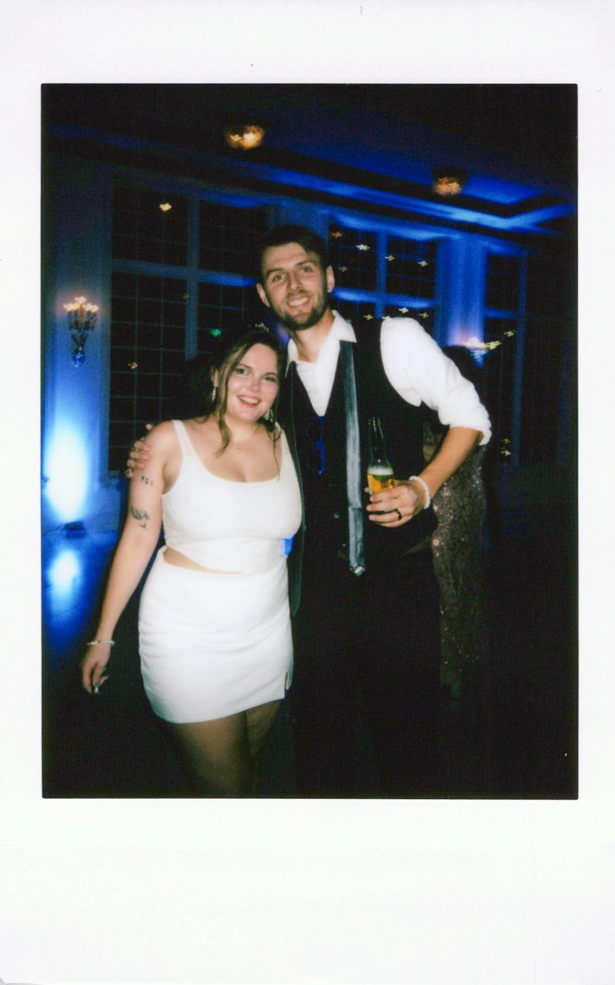 A woman in a white dress and a man in a vest and white shirt are smiling and posing together at an indoor event, with the man holding a drink.