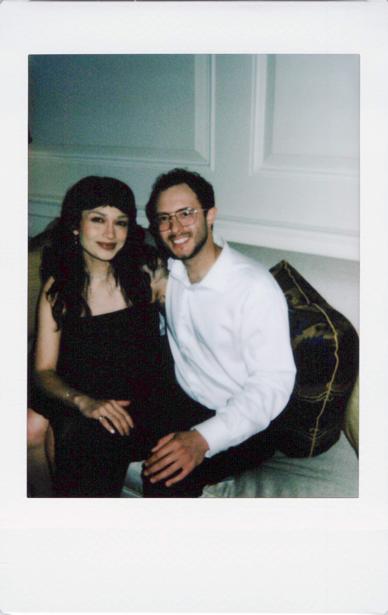 A smiling man with glasses and a woman with dark hair sitting close together, holding hands, inside a room with white paneled walls.