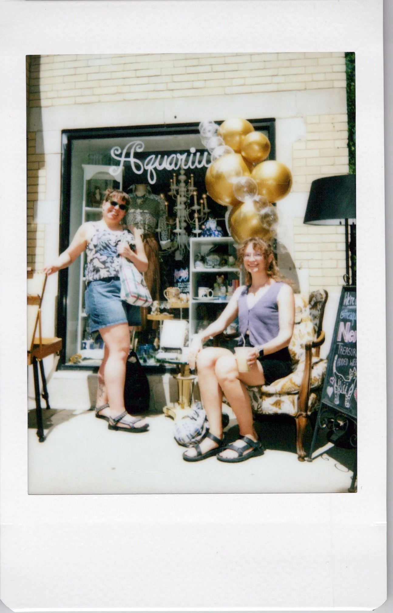 Two women in front of a store window decorated with gold and clear balloons, a sign that says 'Aquarium', and a chalkboard sign outside. One woman is sitting on a vintage chair holding a beverage, and the other woman is standing next to her. Both are smiling.