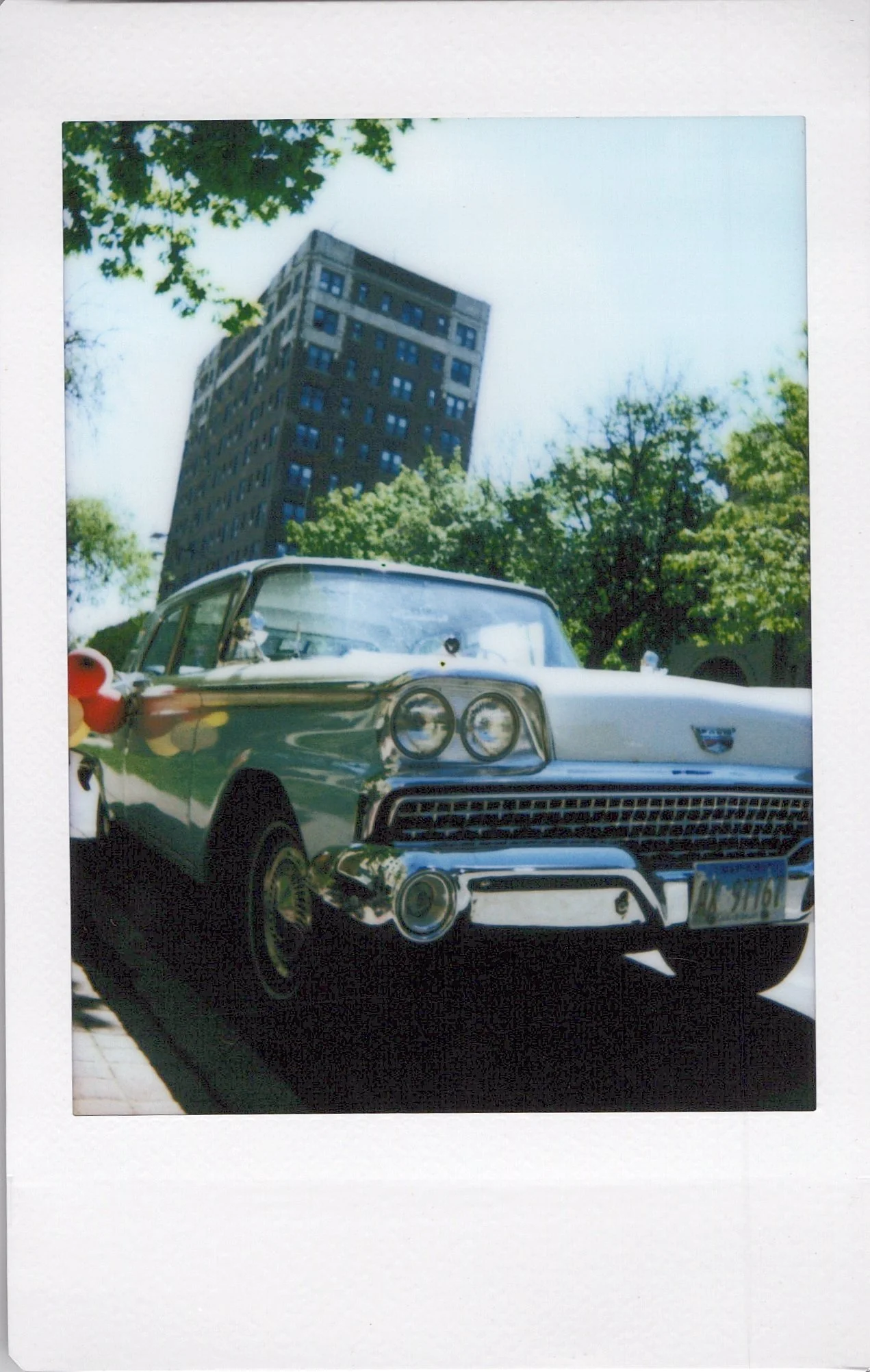 A vintage car parked under a tree with a tall building in the background during daytime.