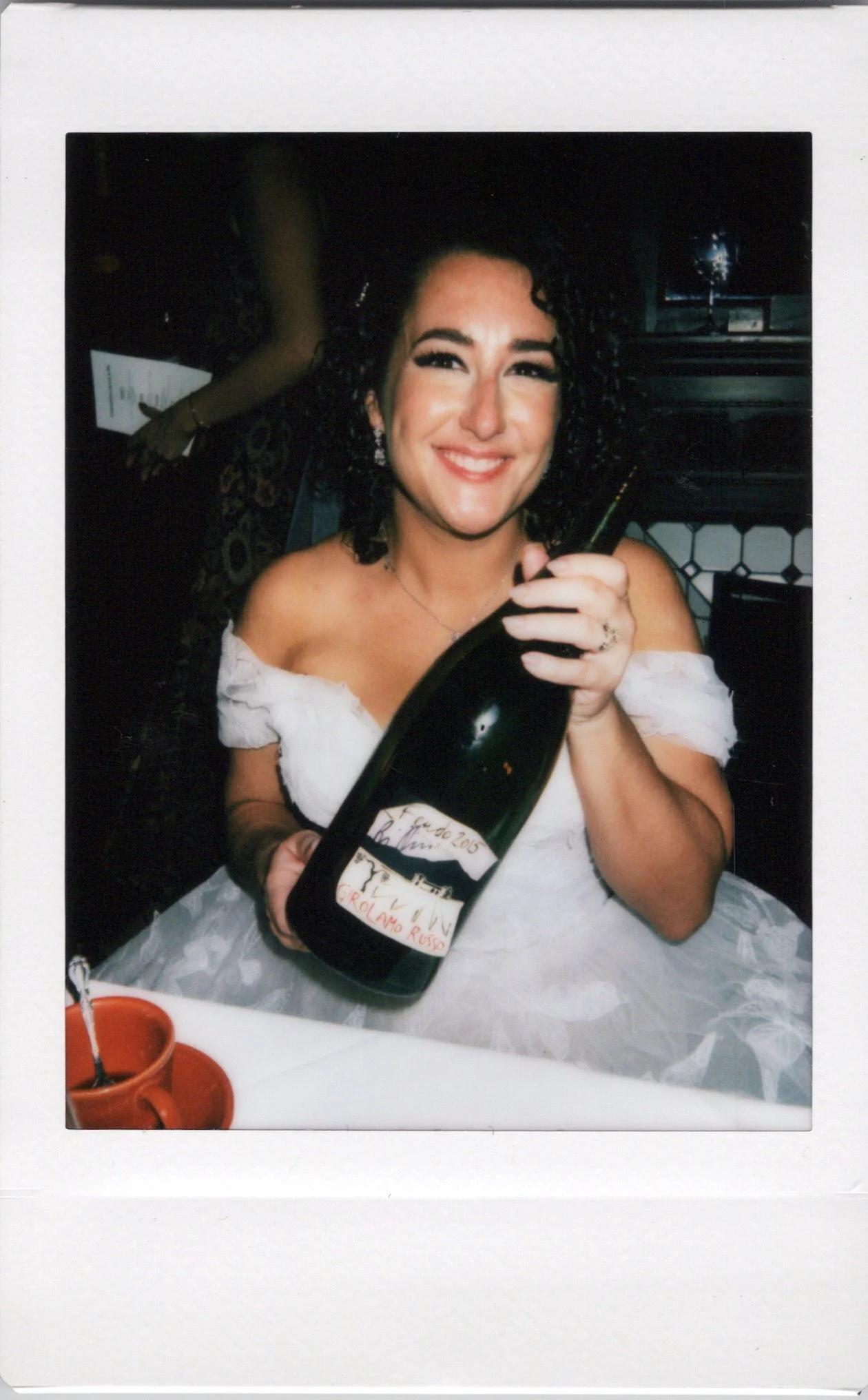 An instant polaroid picture of  smiling bride with curly black hair dressed in a white off-shoulder dress, holding a large wine bottle at a table in Chicago.