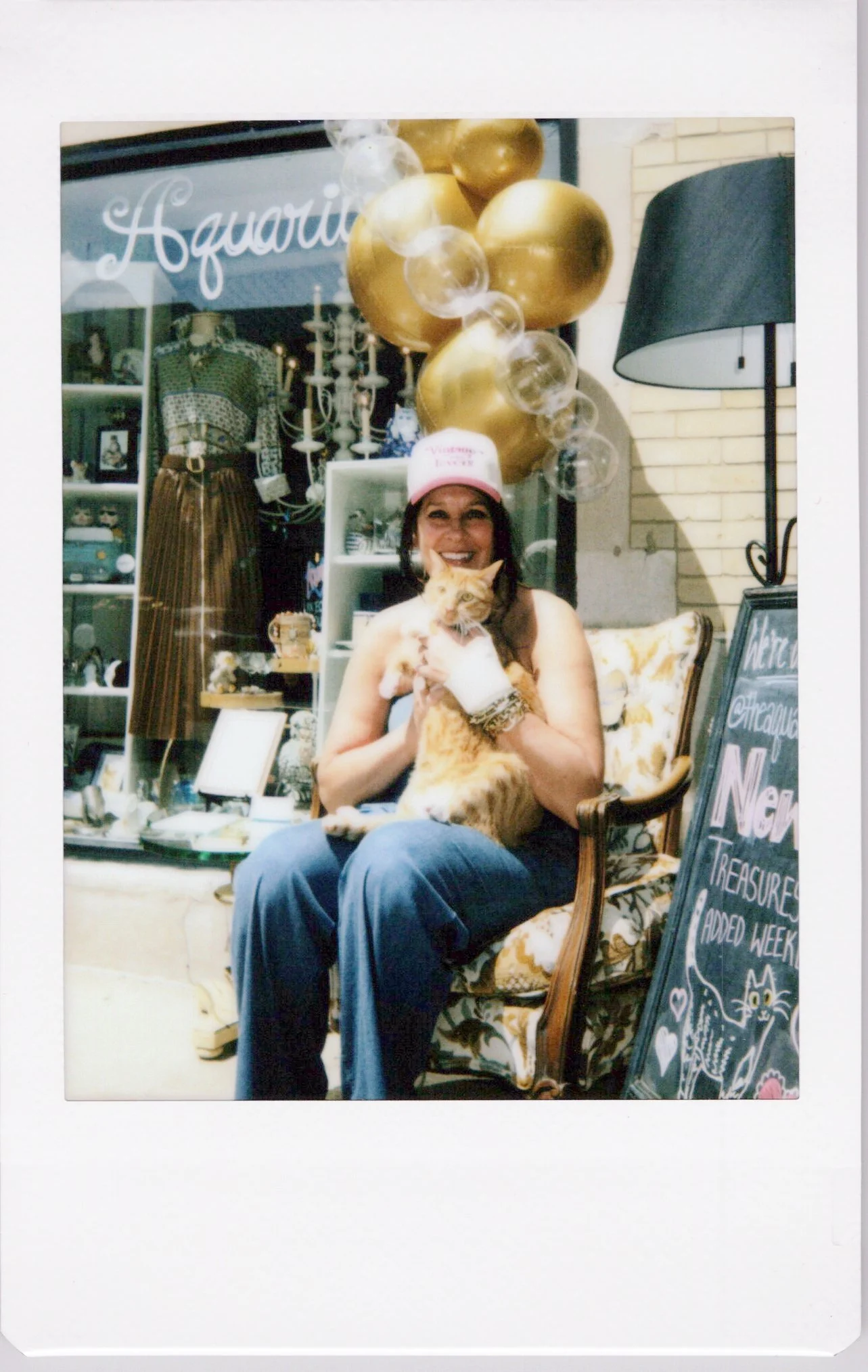 A woman sitting in a vintage armchair holding an orange tabby cat, with balloons and store display in the background.