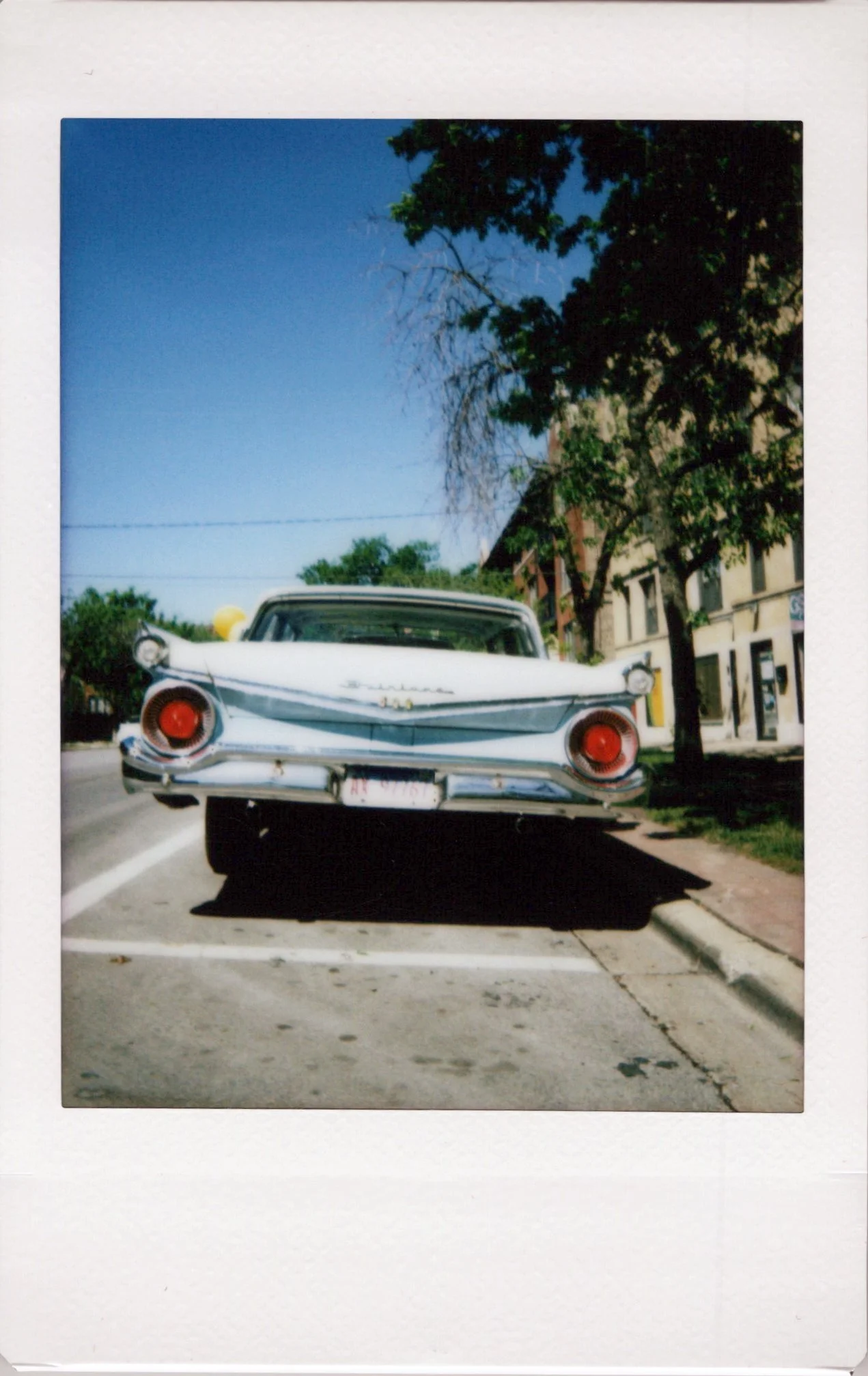 Color photograph of a vintage white Cadillac car parked on a city street with buildings and trees in the background, under clear blue sky.