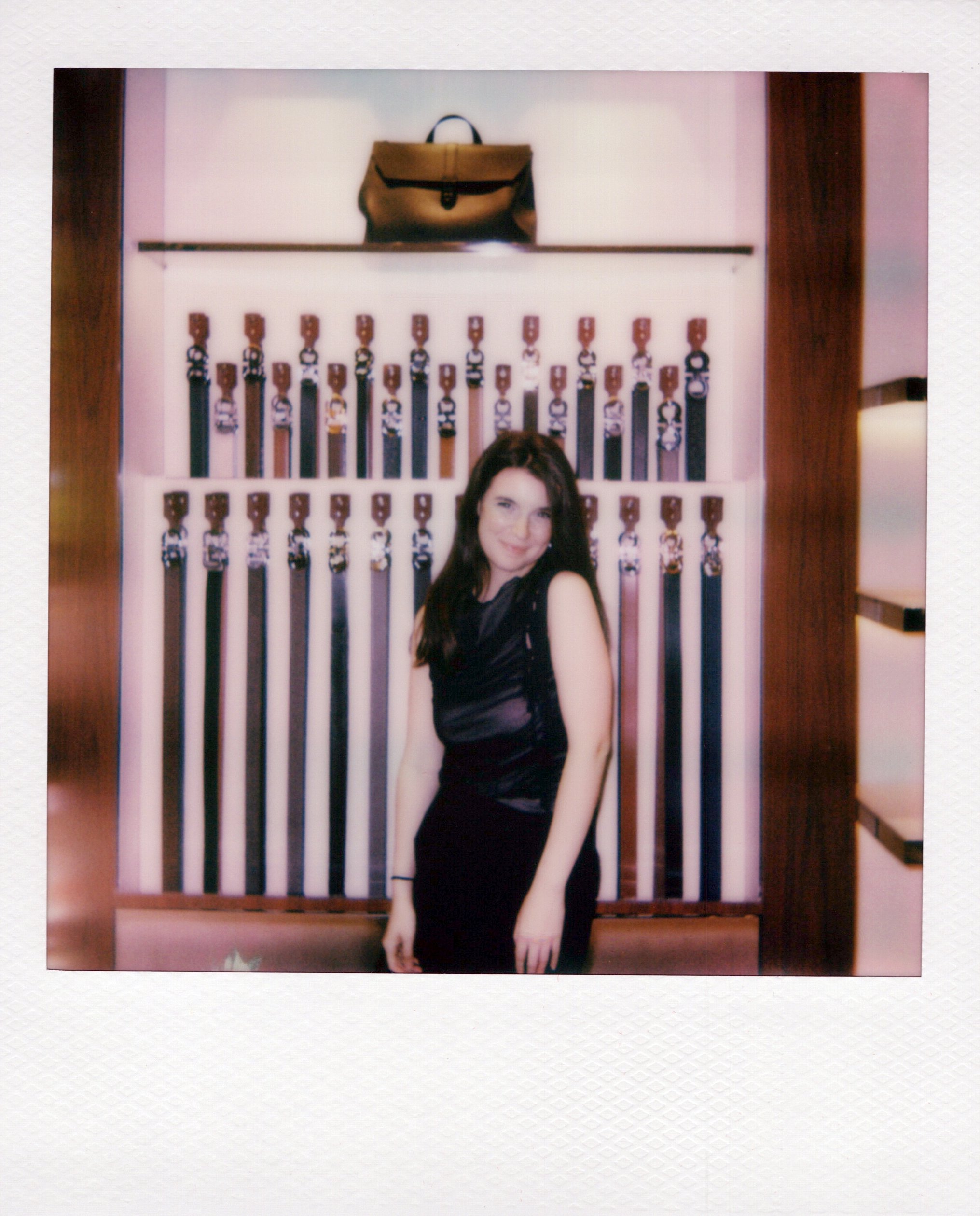 A woman with long brown hair in a black dress standing in front of a wall display with rows of hanging ribbons or medals and a brown handbag on a glass shelf above the display. At ferragamo chicago