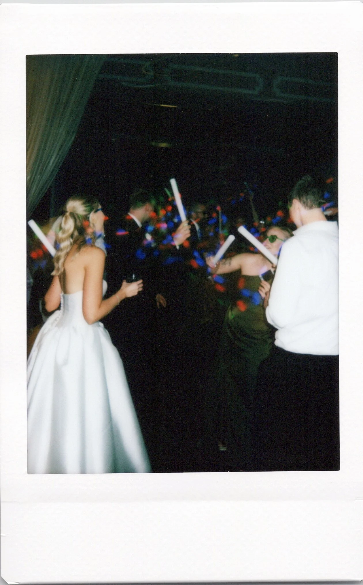 People dancing at a party with glow sticks and colored lights, including a woman in a white dress and others in formal attire.