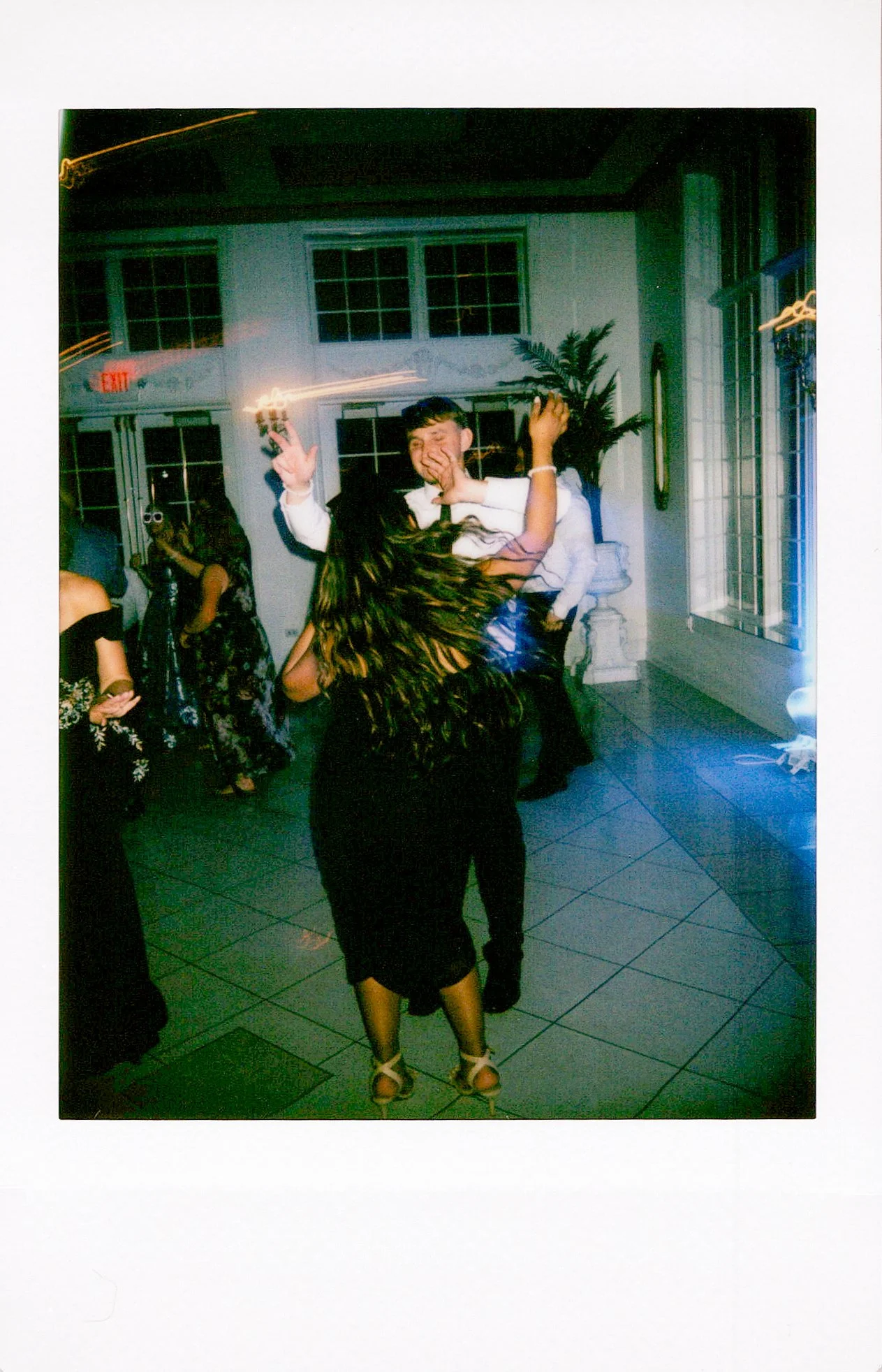 People dancing and socializing indoors with tiled floor, white walls, large windows, and plants, at an event or celebration.