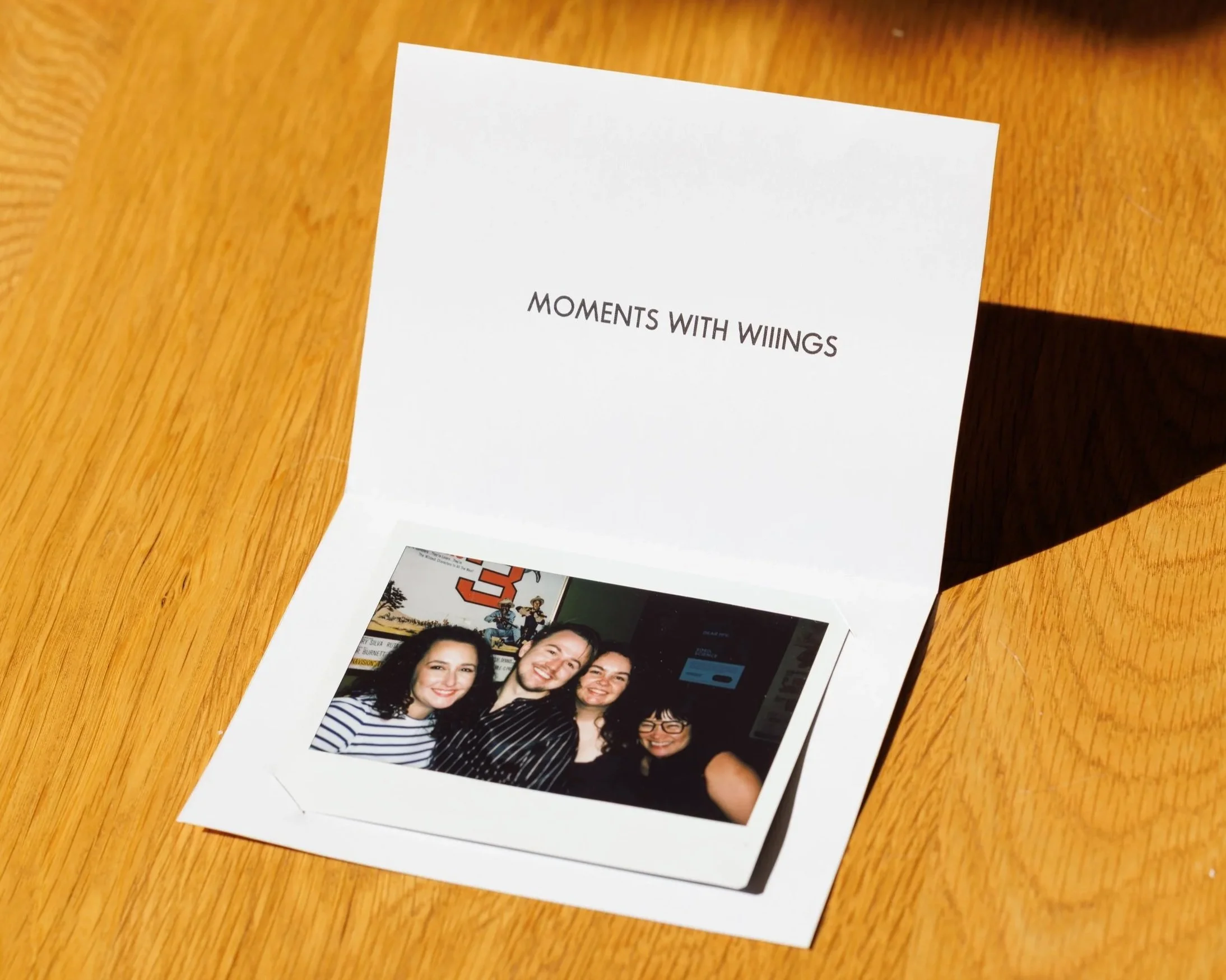 Photograph of a group of four women smiling and posing together for a selfie, with a background of a wall with various posters and a chess piece. The photo is printed on a glossy photo paper.