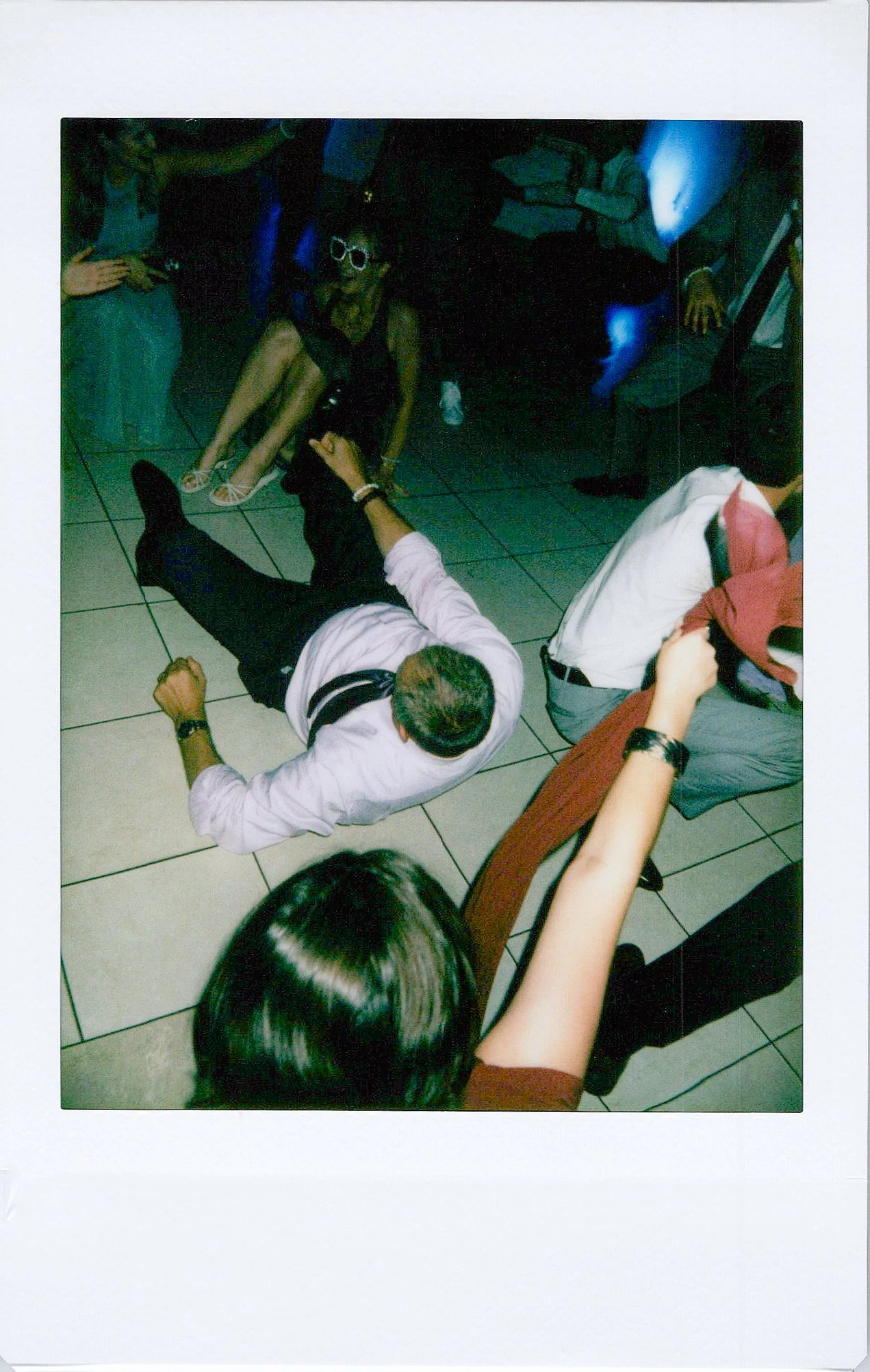 People sitting on the floor and leaning against chairs in a dimly lit room, some taking photos or videos with their phones.