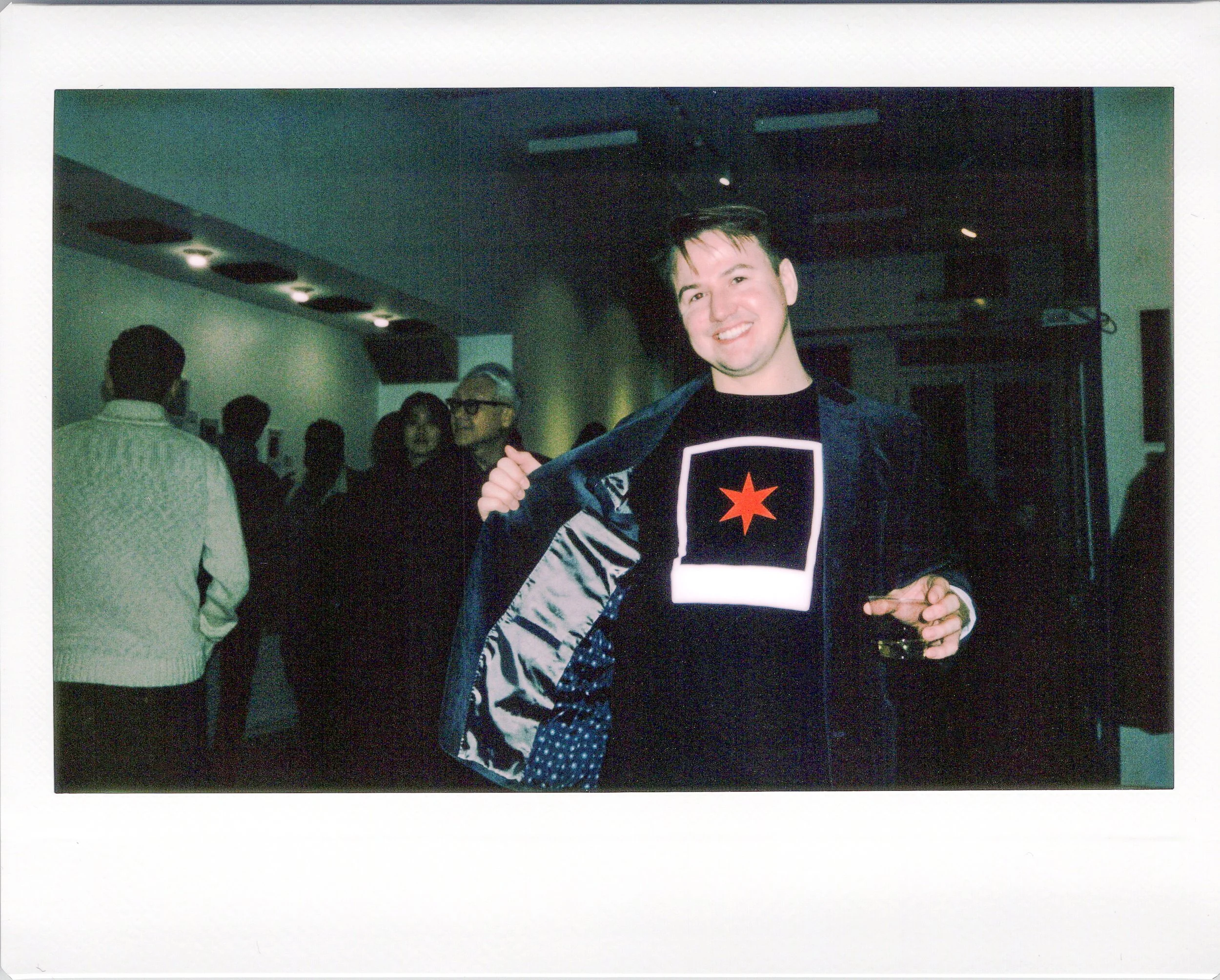A young man with short dark hair, smiling and wearing a black t-shirt with a chicago red star design, is opening a dark blue jacket with a shiny inner lining in a crowded chicago gallery.