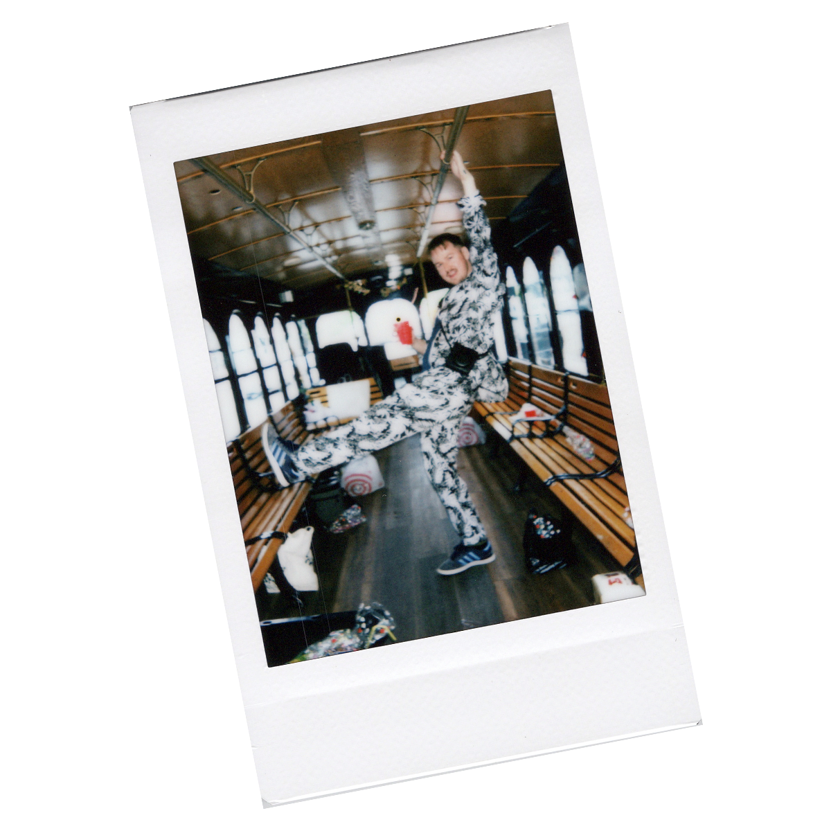 A person in a black and white patterned jumpsuit hanging from a pole inside a vintage bus with wooden benches and large windows.