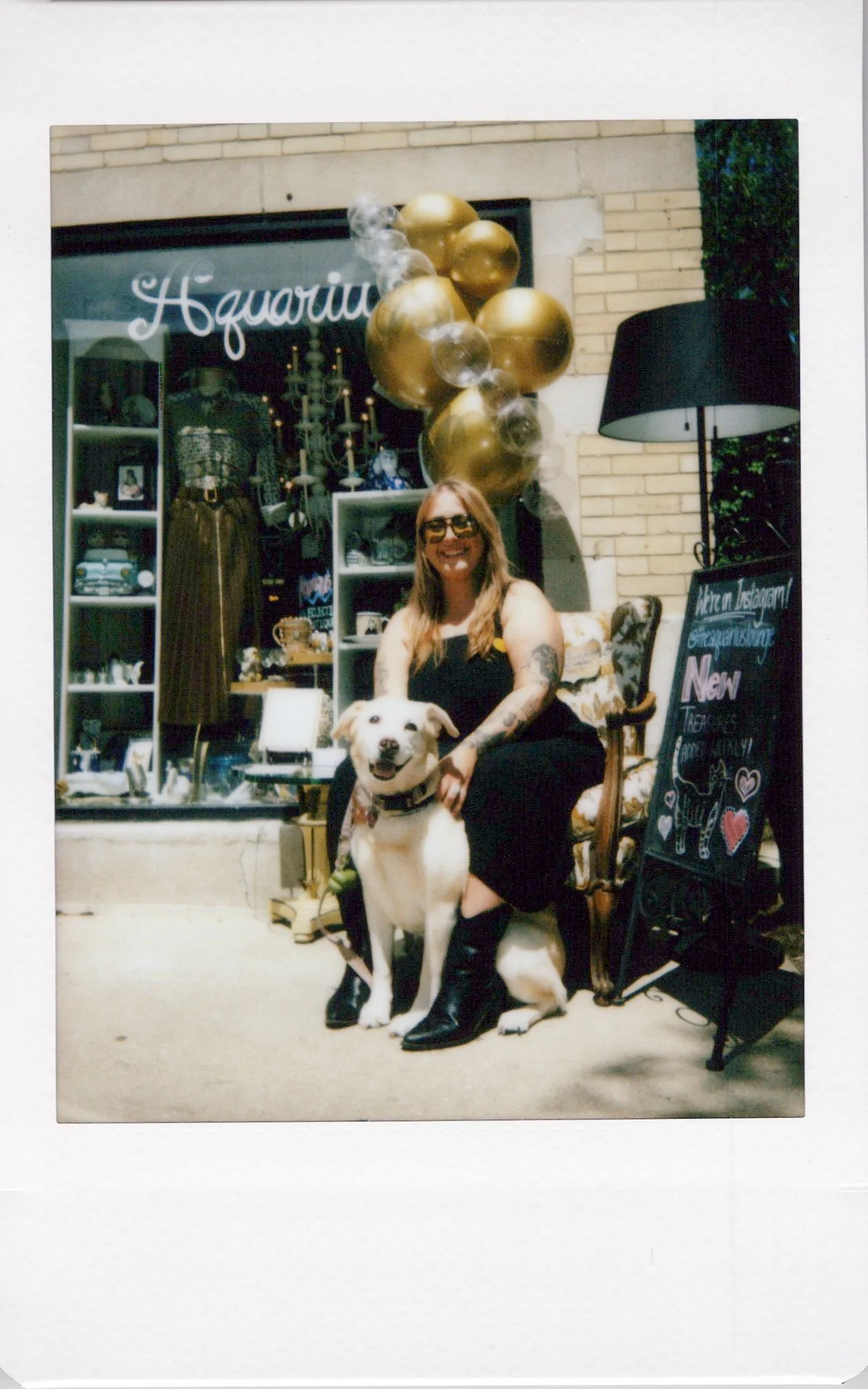 A woman with tattoos and sunglasses sitting on a vintage chair, smiling and holding a dog, in front of a storefront decorated with gold balloons, candles, and a chalkboard sign.