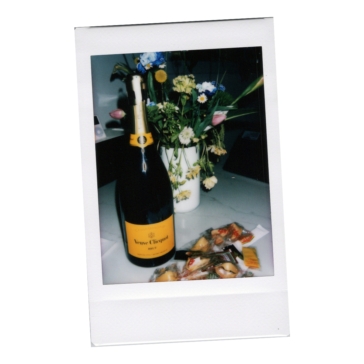 A Polaroid-style photo depicting a bouquet of flowers in a white vase, a bottle of Veuve Clicquot champagne, and some snacks on a table.