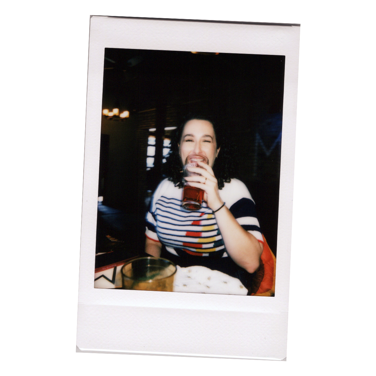 A woman with curly dark hair, wearing a striped shirt, laughing and holding a drink inside a dimly lit restaurant or bar.