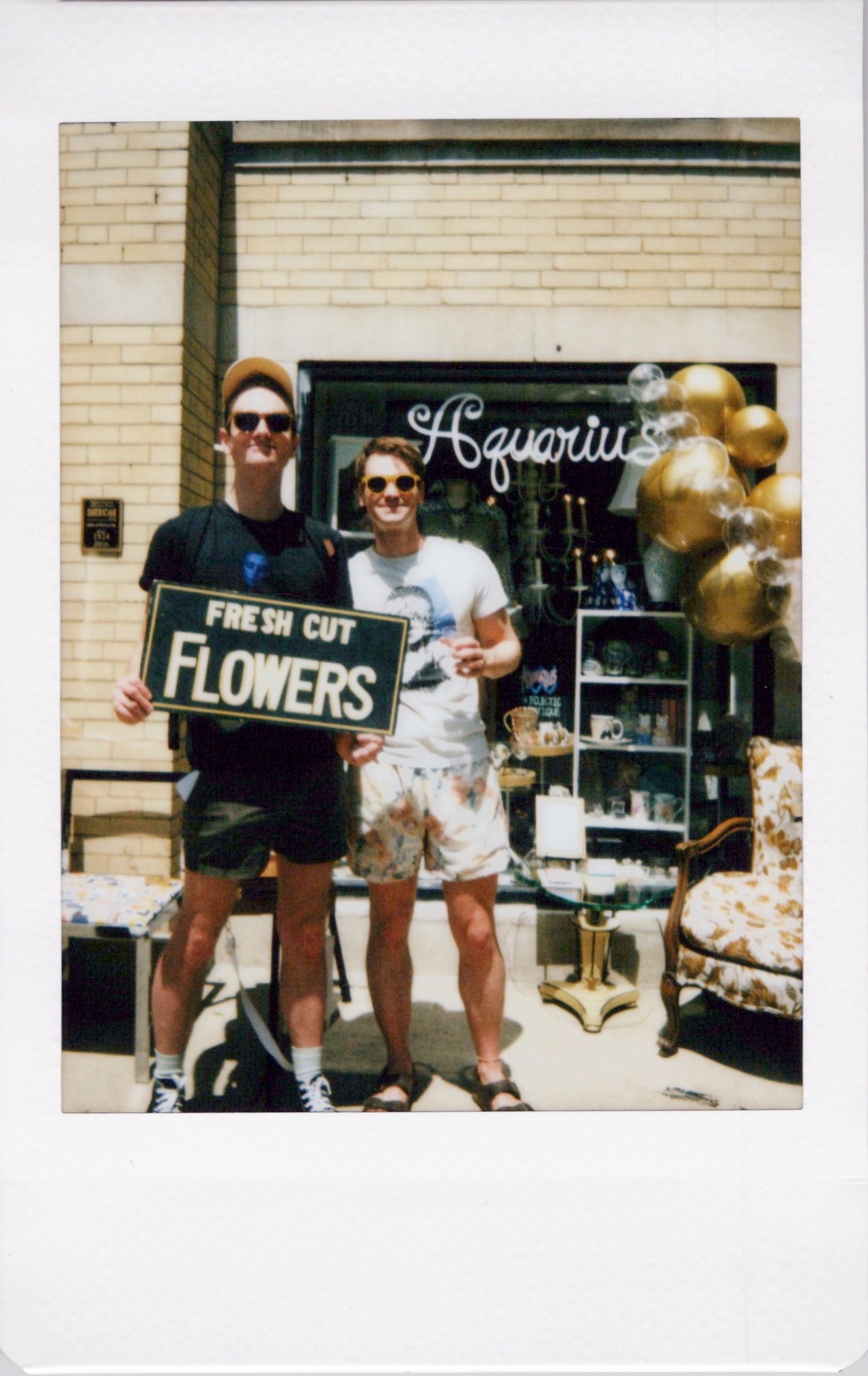Two young men standing outdoors in front of a shop window, one holding a sign that says "Fresh Cut Flowers." They are wearing casual summer clothing, sunglasses, and standing on a sidewalk with furniture and balloons nearby. Chicago on-site photo