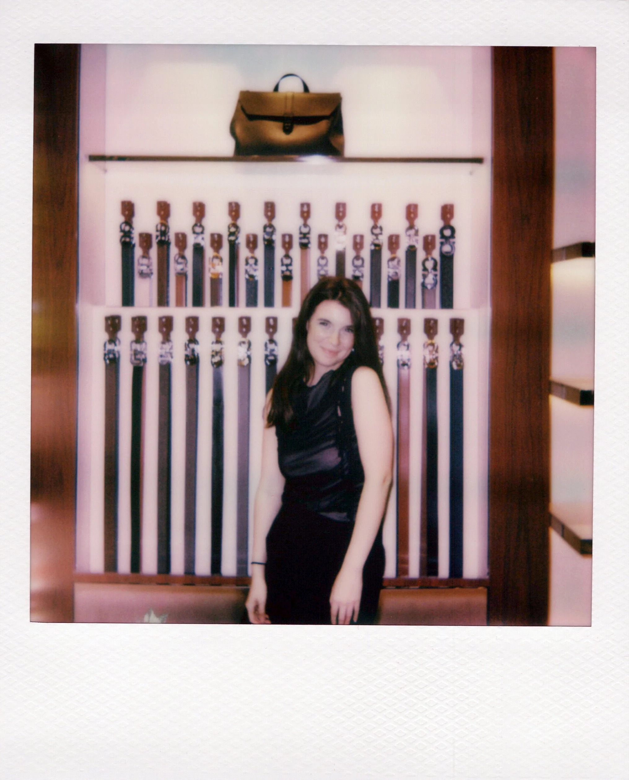 A woman with long dark hair wearing a black dress, standing in front of a wall decorated with multiple rows of belts and a brown bag on a shelf above the belts at Ferragamo Michigan Ave.