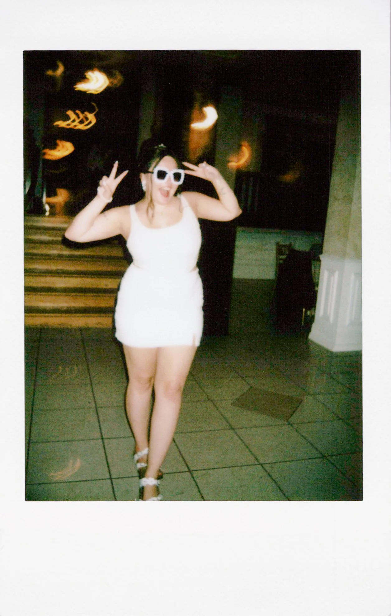 A woman in a white dress and sandals posing with peace signs, wearing large white sunglasses, standing indoors with a dark background and stairs.