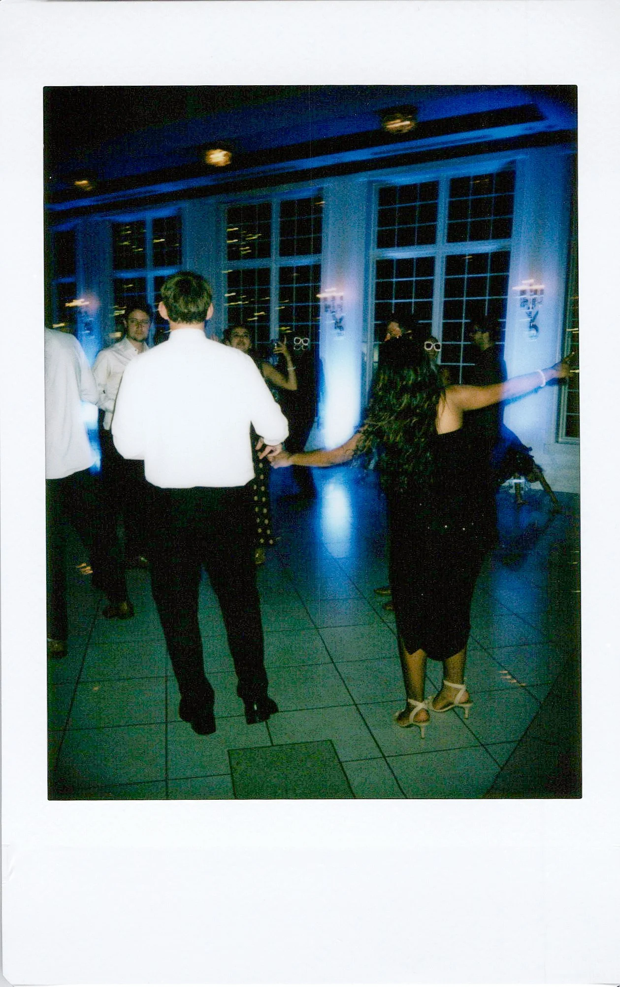 People dancing at a party with blue lighting in a large room with tall windows.