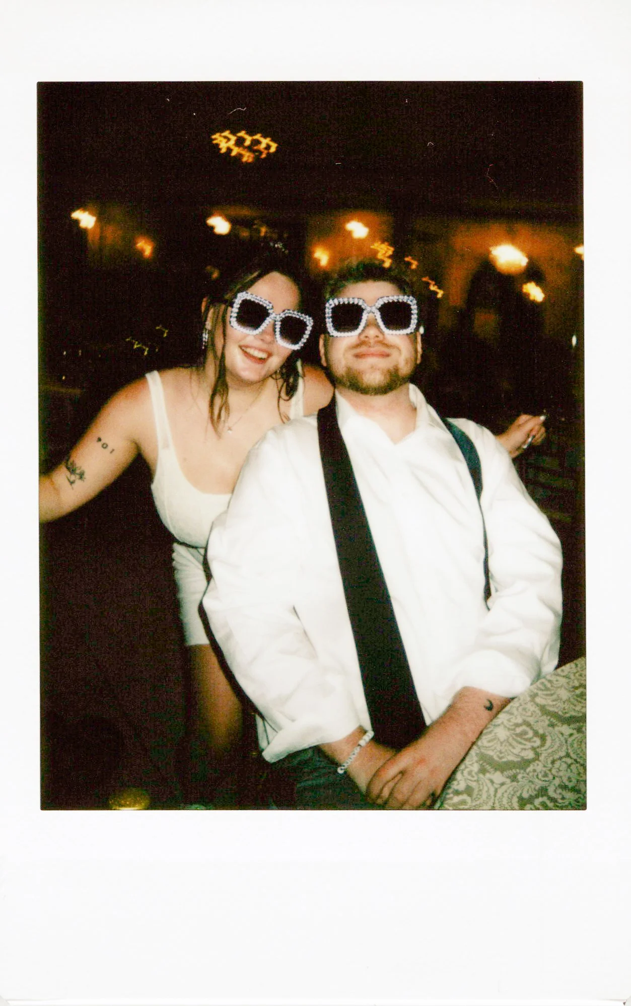 Two people wearing large, sparkly sunglasses at a party or celebration, smiling and posing for the camera, with warm yellow lights in the background.