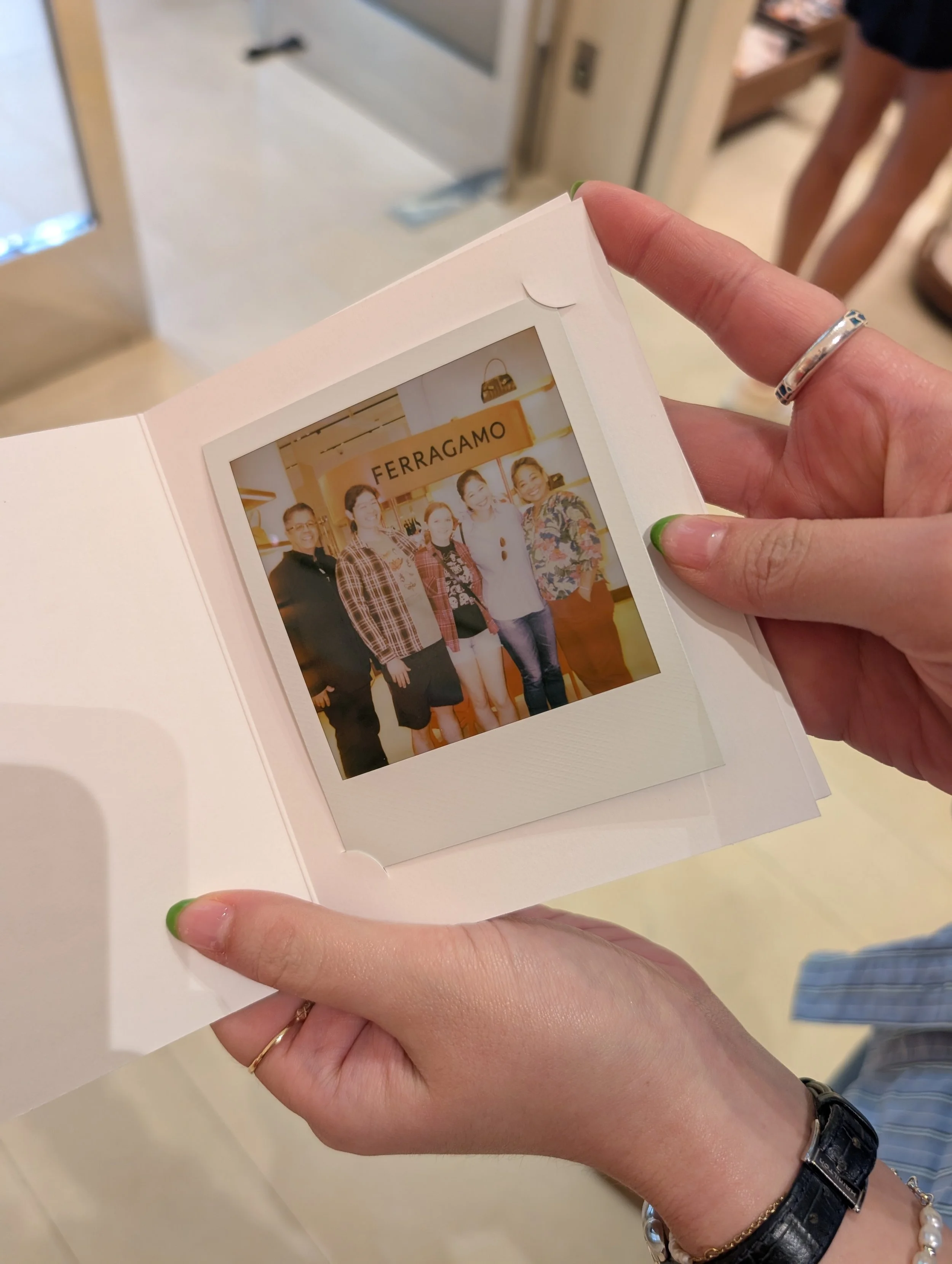 A person holding a picture book showing a photo of five women standing in front of a store with a sign that reads 'FERRAGAMO'. The person has a silver ring on their finger and is wearing a black watch and pearl bracelet. The background shows a person in shorts and sandals, and an elevator door.