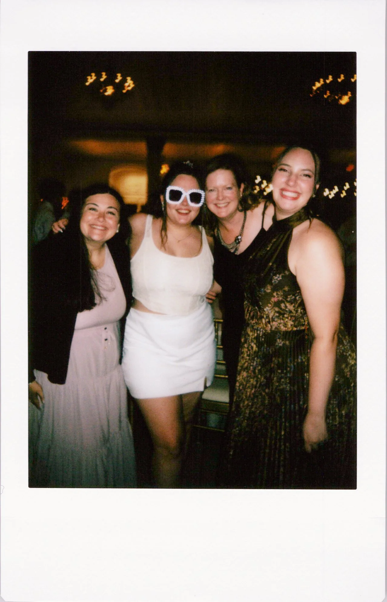 Four women smiling and posing together indoors at a party or celebration, with warm lighting and blurred background.