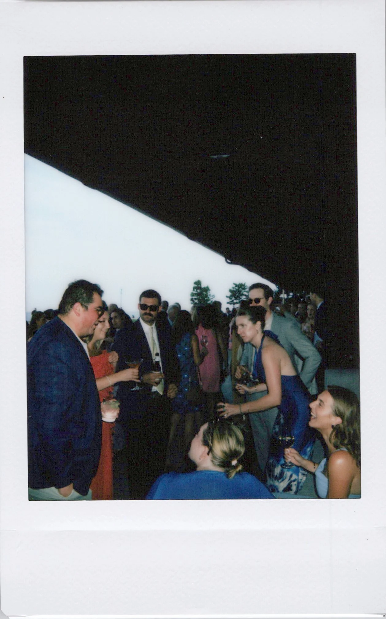 People socializing at an outdoor event, some holding drinks, with a tent overhead, outdoors during the daytime.