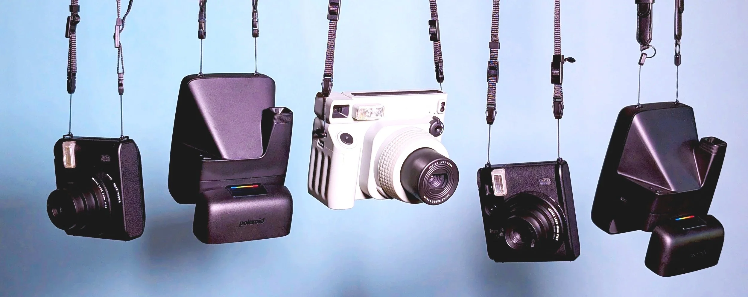 A collection of instant cameras hanging from their straps with a blue background. Instax mini 99, Polaroid Flip, Instax Wide 400