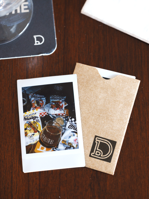 A photo of a cardboard folder with a 'D' logo on a wooden table, next to a Polaroid picture showing a Chicago sports memorabilia collection, including a mug and various patches.