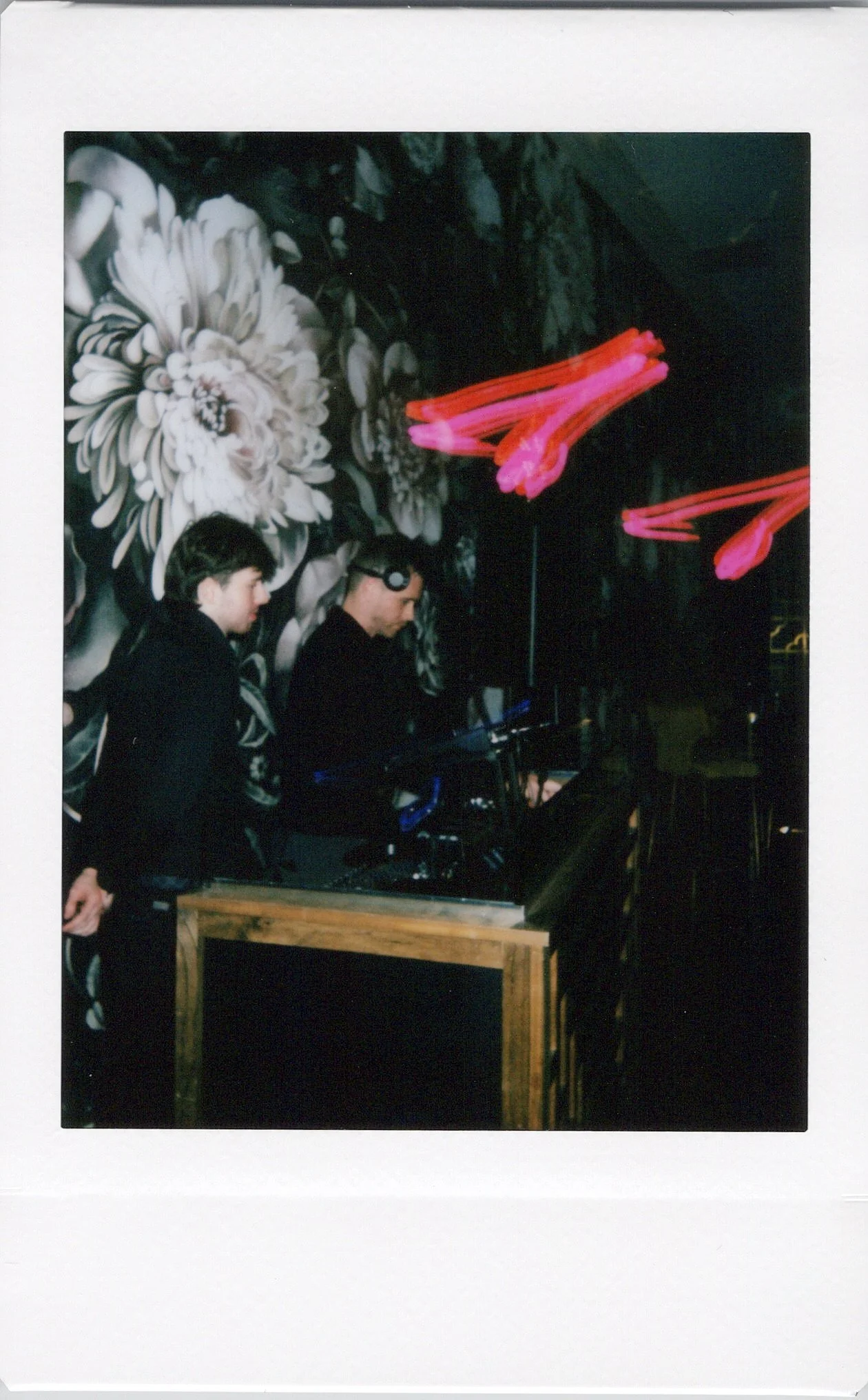 Two men stand behind a DJ setup in a dimly lit venue with a floral mural background and neon light decorations.
