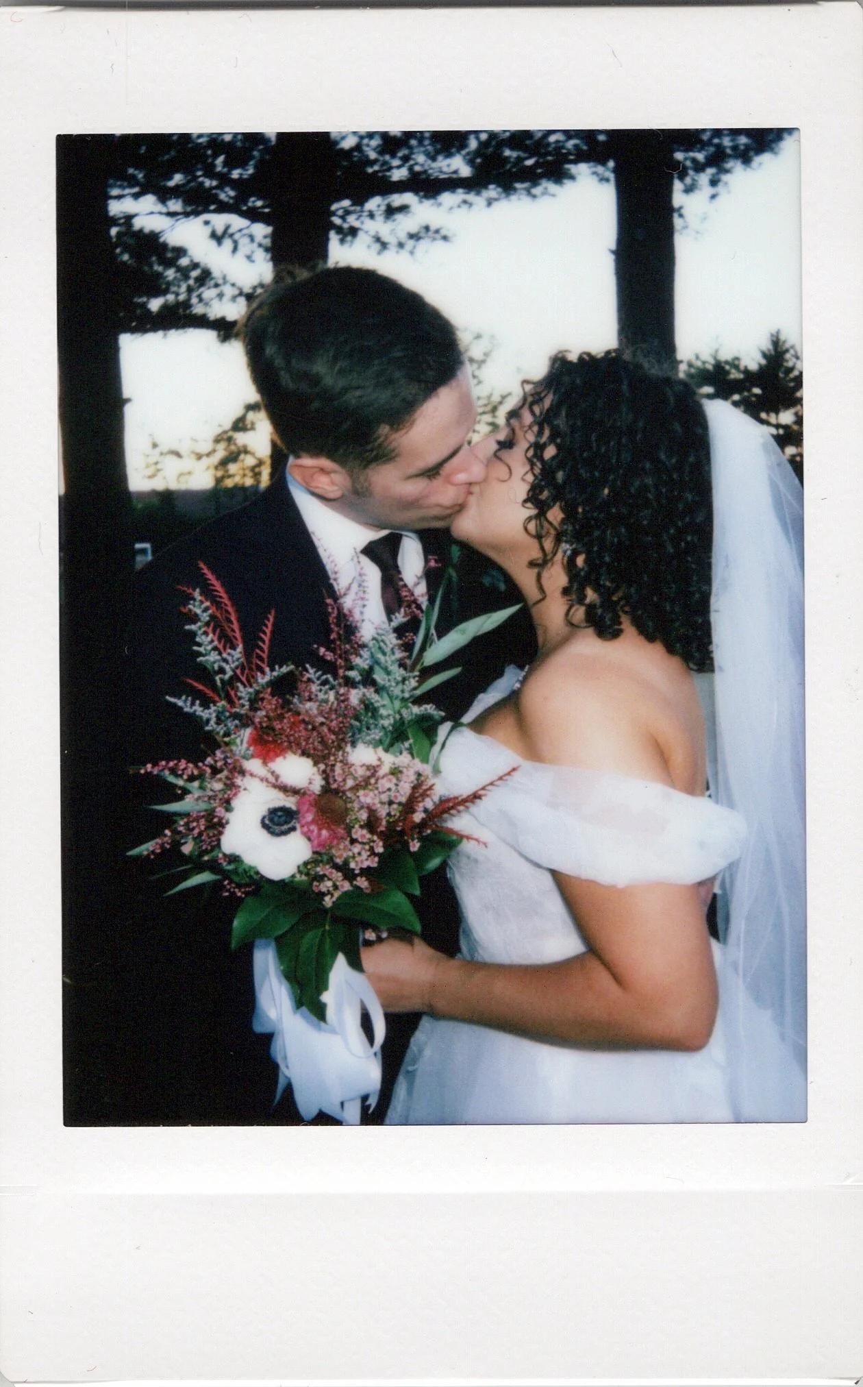 A bride and groom sharing a kiss outdoors during their wedding, with trees in the background and the bride holding a bouquet of flowers. Photo booth chicago polaroid wedding