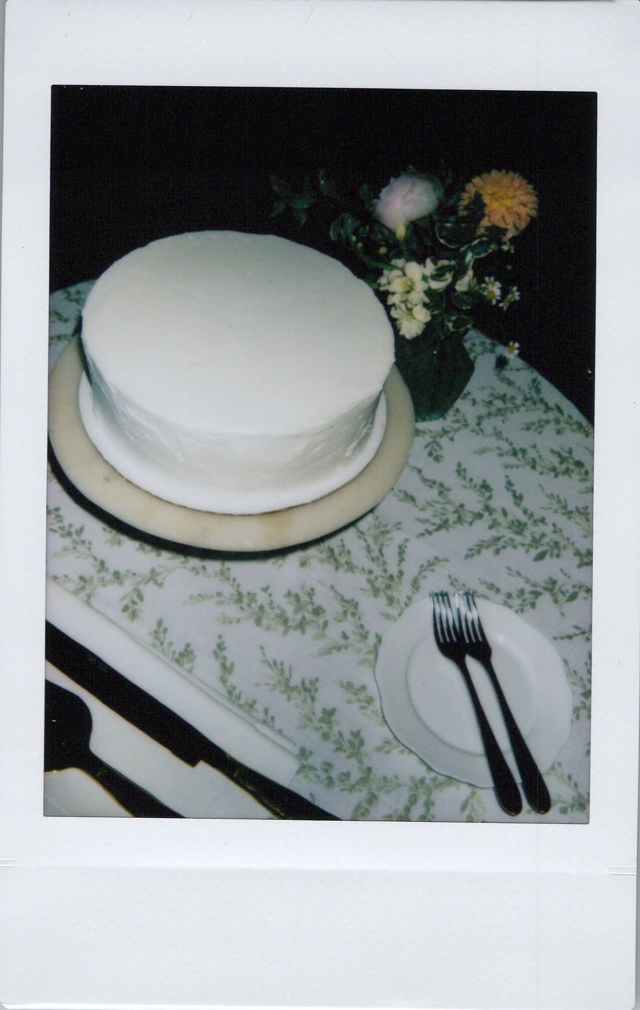 A cake on a round plate and a floral centerpiece with white, pink, and orange flowers. The cake is white and round, placed on a table with a patterned tablecloth. There is a fork and a knife on a small white plate nearby.