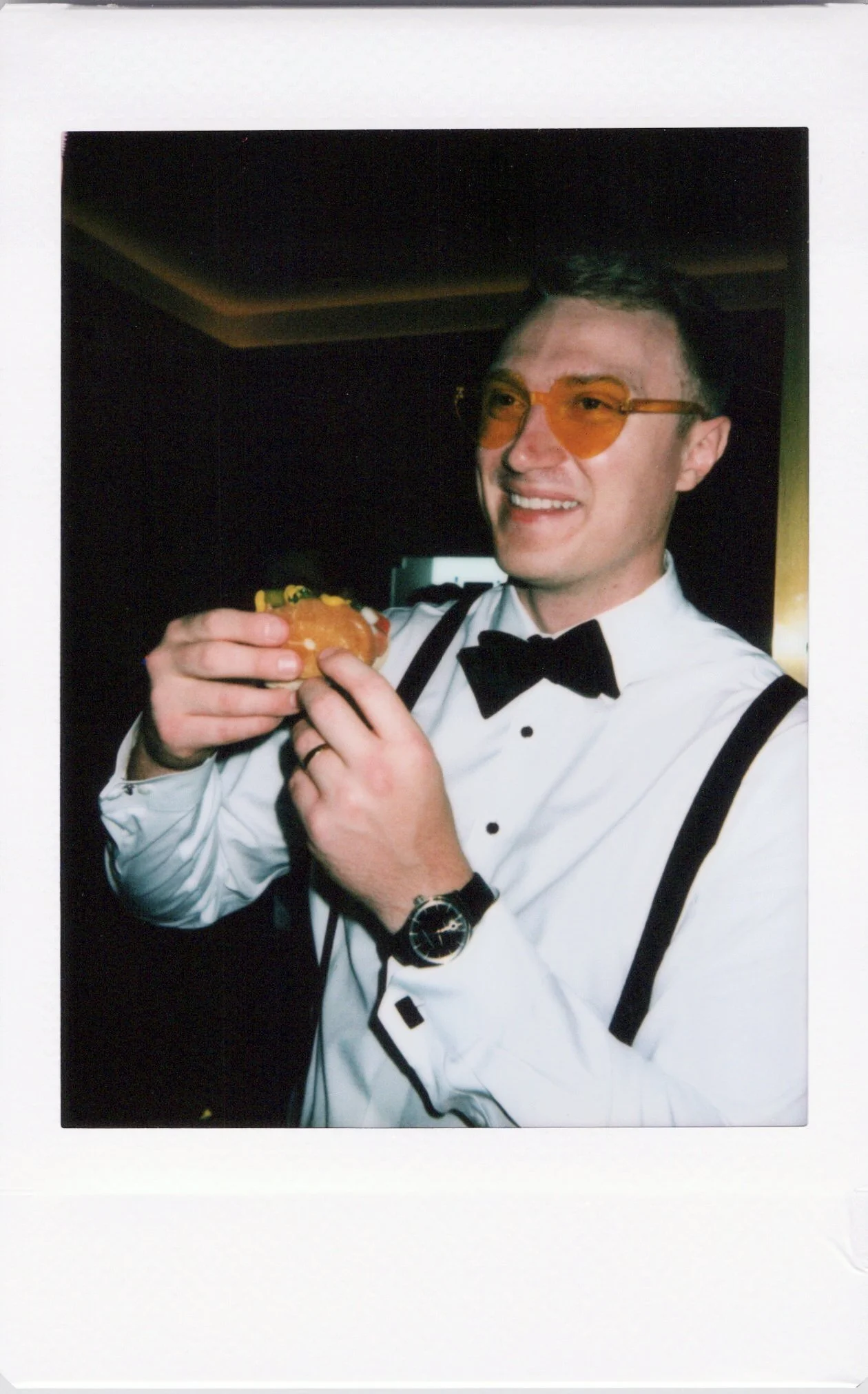 A groom wearing a white dress shirt, black bow tie, black suspenders, orange-tinted glasses, and a wristwatch, holding a hot dog and smiling.