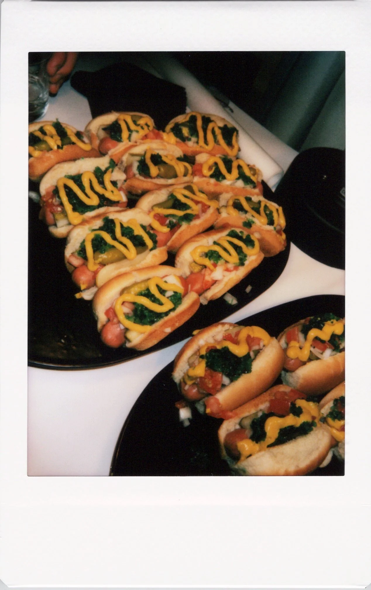 A polaroid instant photo of Chicago dogs topped with mustard and relish on black plates at a wedding.