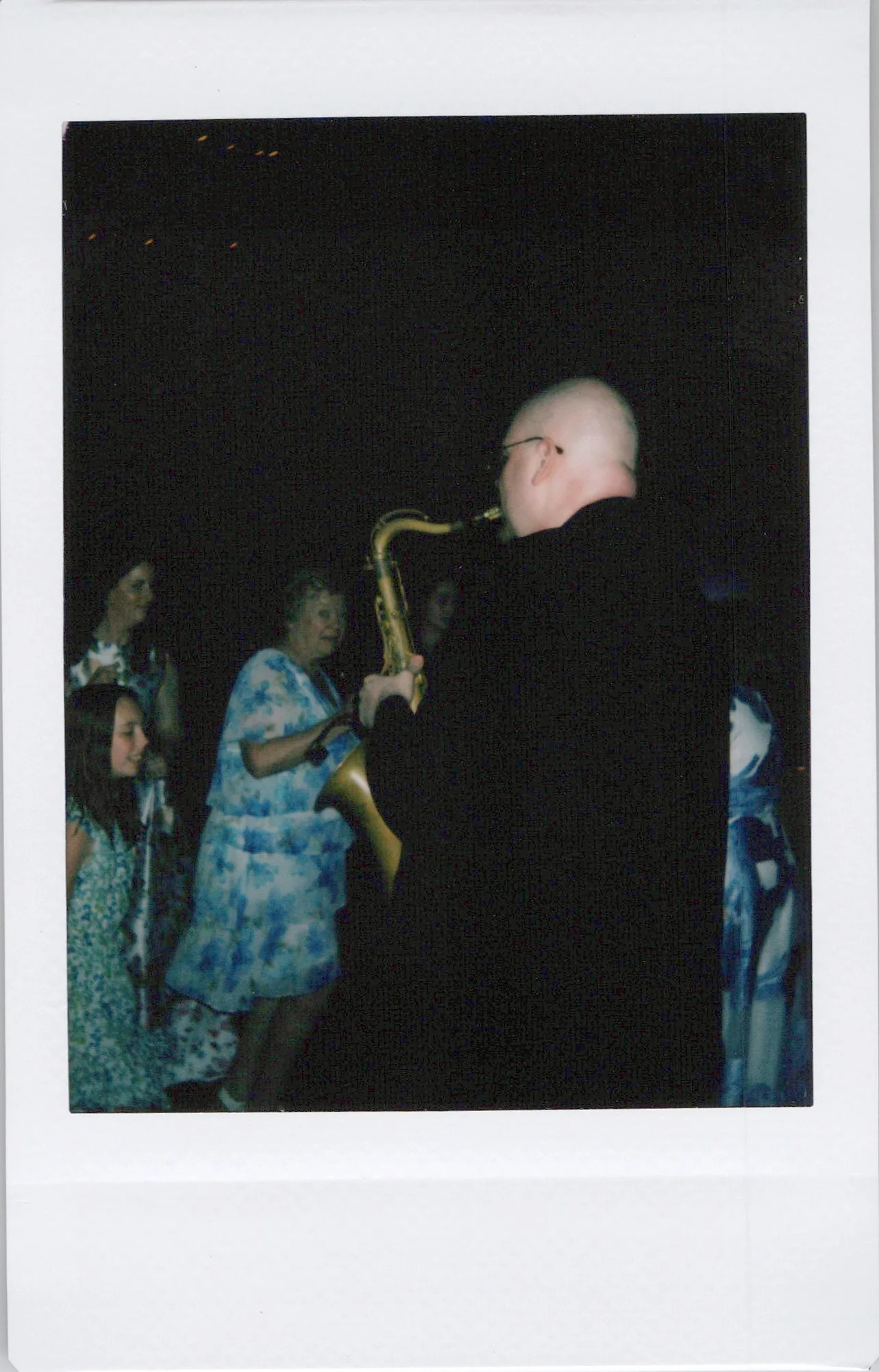 A man with a shaved head playing a saxophone at a night event with women wearing matching blue and white patterned dresses surrounding him.