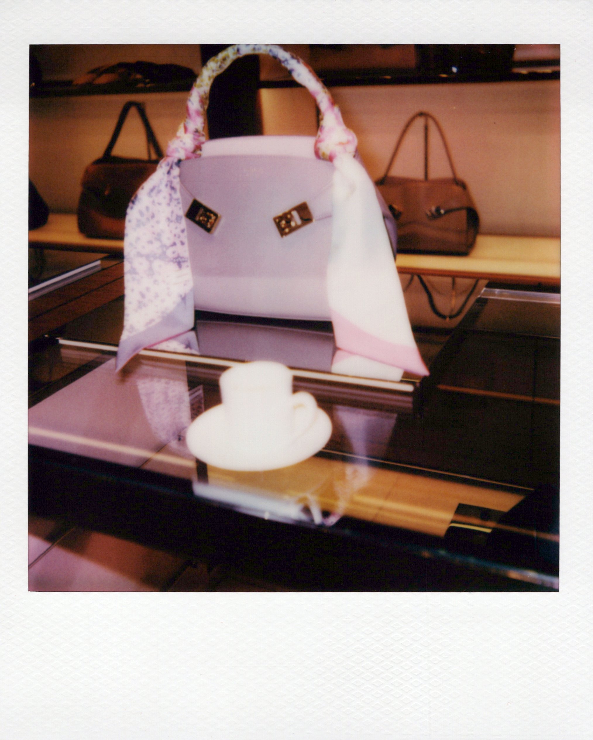 A polaroid of a white handbag with gold hardware and a floral scarf tied around the handle, placed on a table with other handbags in the background at a brand activation in Chicago.
