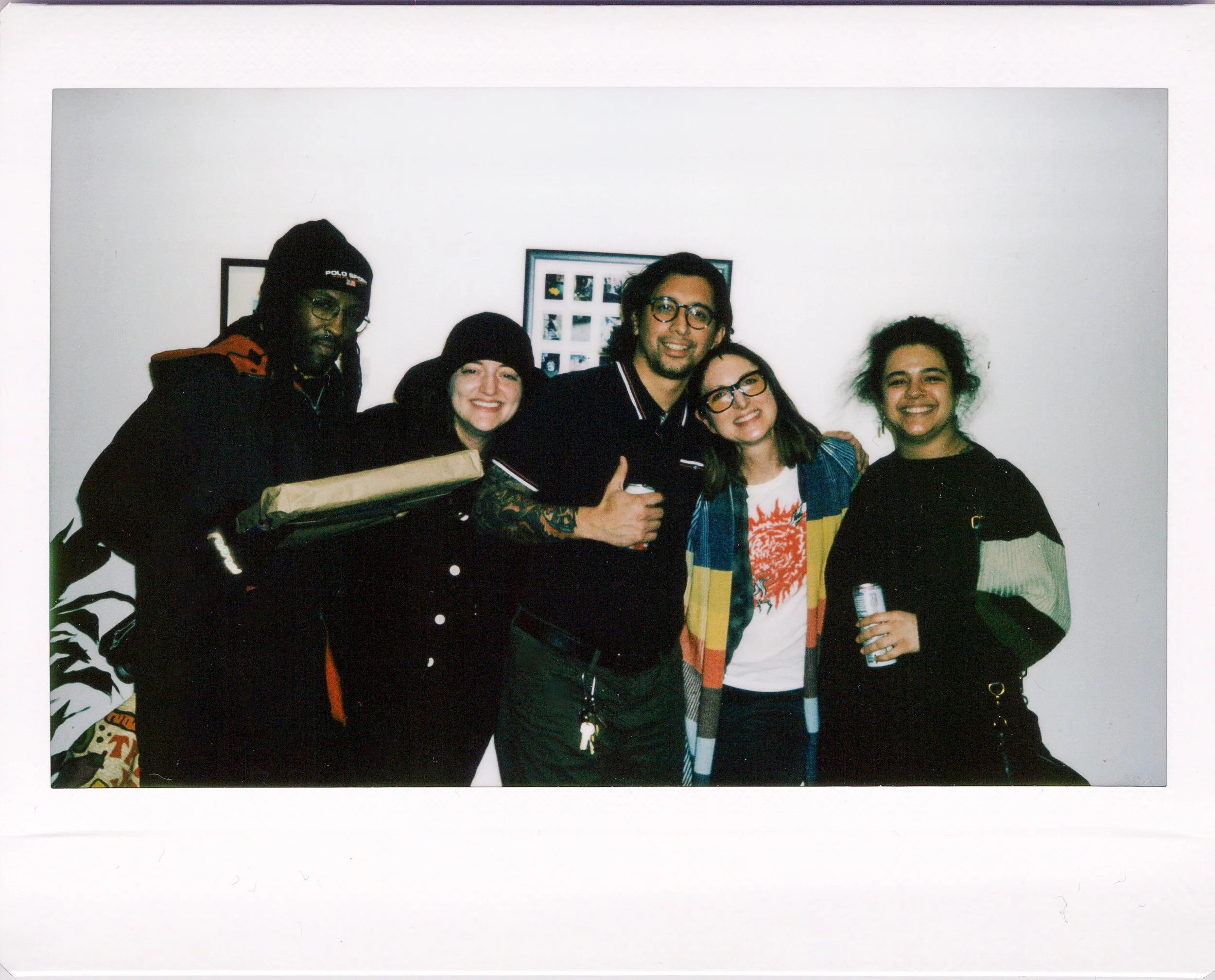A group of five young adults standing together and smiling, with a white wall and framed photographs in the background.