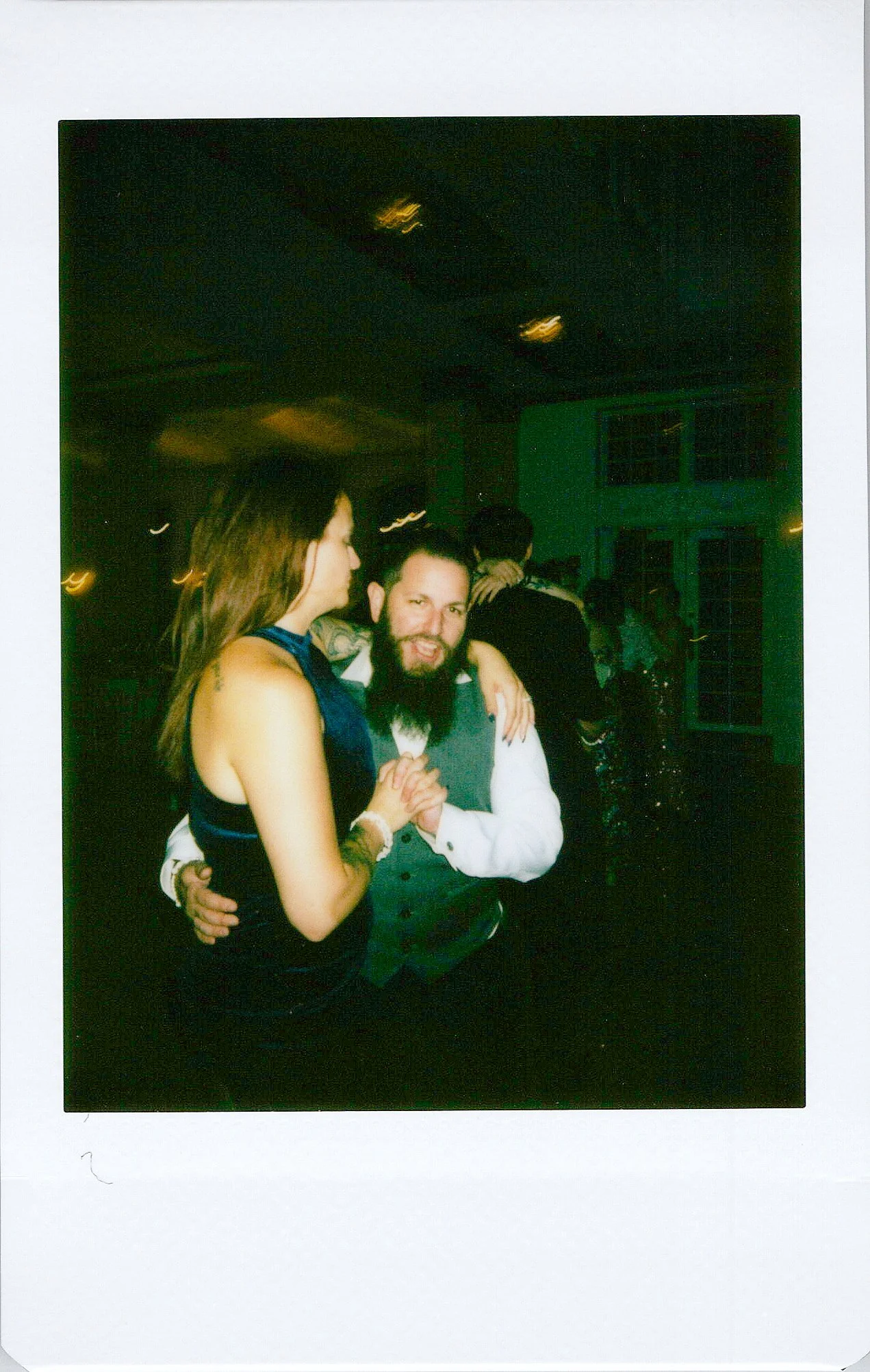 A man and woman dancing closely in a dimly lit room, with a woman in the background wearing dark clothing.