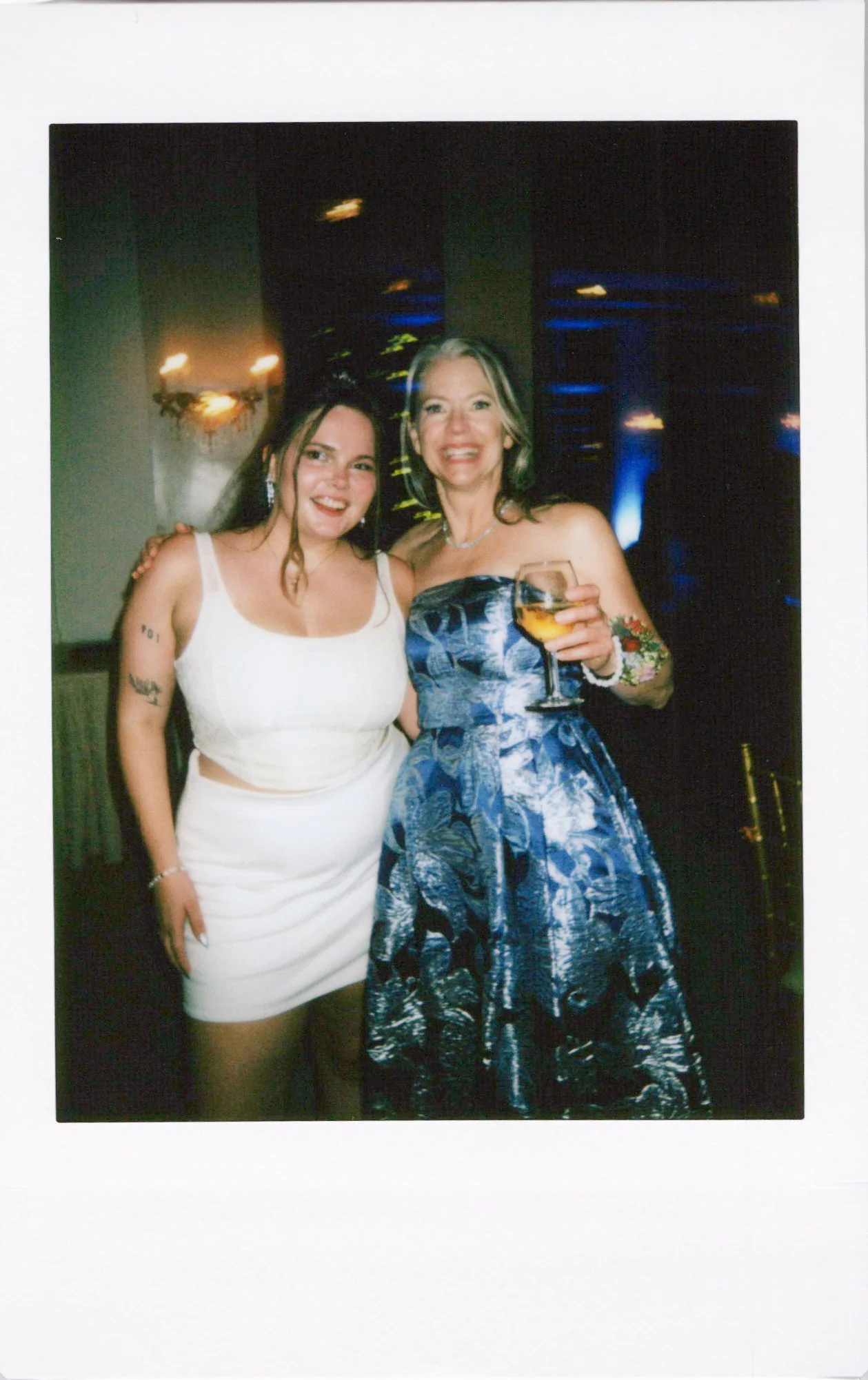 Two women smiling at an event, one in a white dress and the other in a blue patterned strapless gown, holding a glass of wine, with a dark background and ambient lighting.