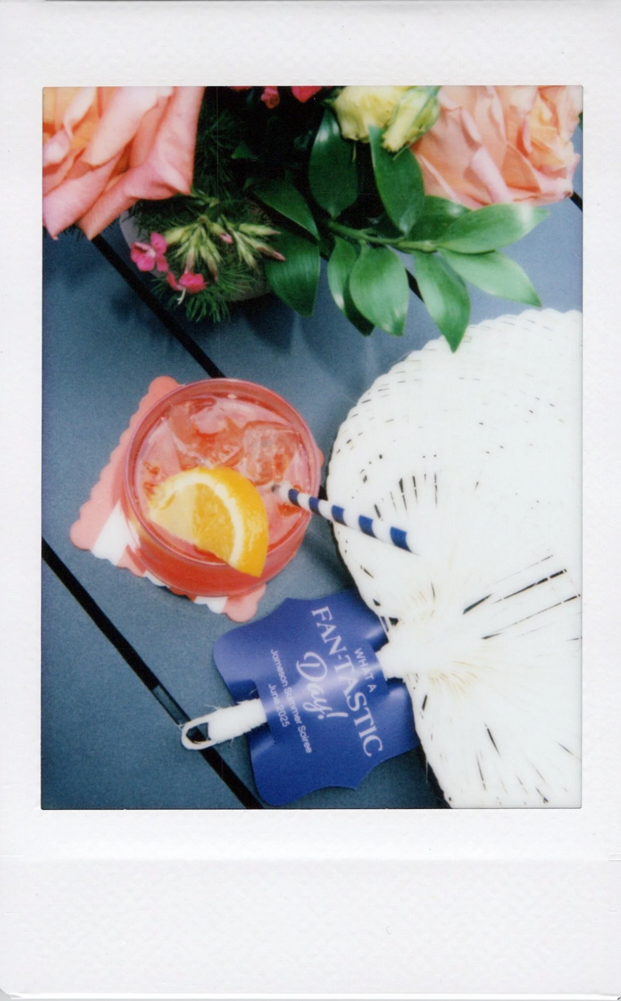 A bouquet of pink roses and green leaves, a pink drink with an orange slice and straw, and a white butterfly-shaped fan with a blue fan-shaped tag on a dark table.