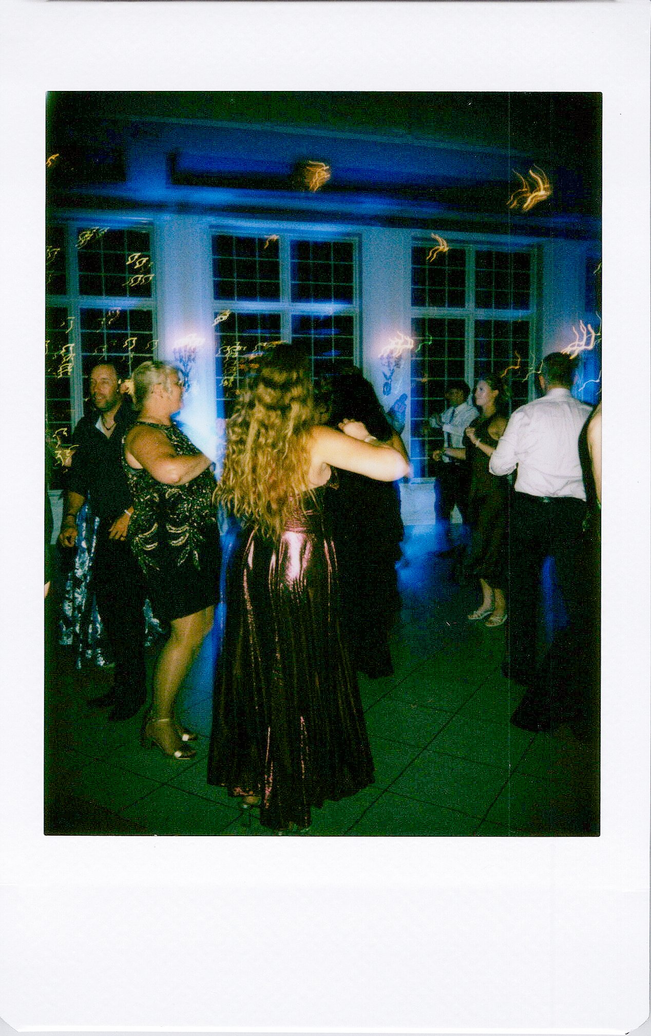 People dancing and socializing at an indoor party with large windows and blue lighting.