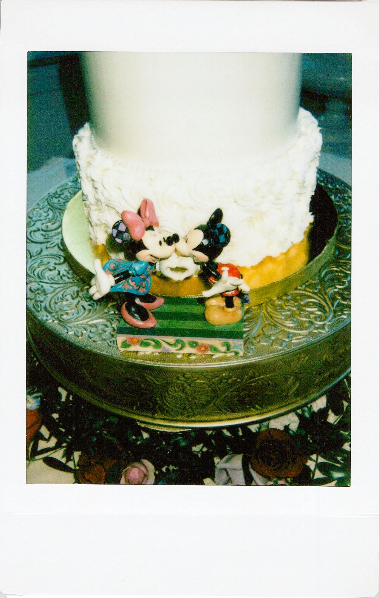 A white cake decorated with two Mickey Mouse and Minnie Mouse figurines at the base, on a decorative golden tray with intricate patterns.