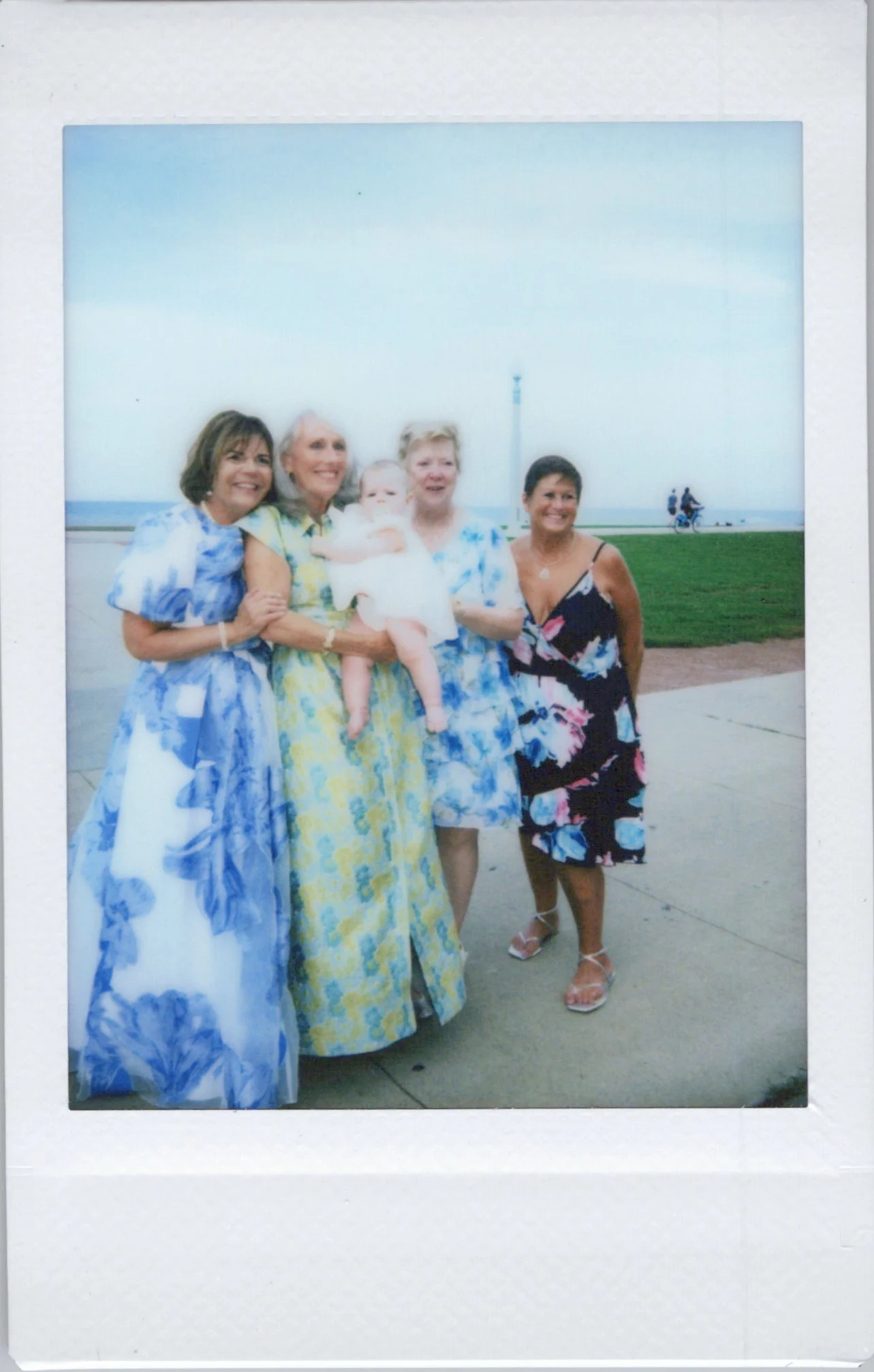 Group of four women and a baby standing outdoors near the beach, dressed in colorful summer dresses, with a grassy area and the ocean in the background.