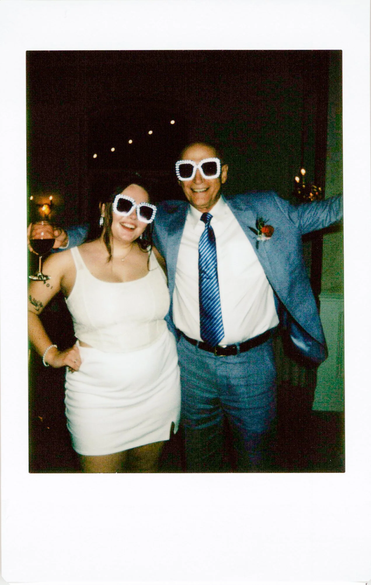 Two people at a celebration wearing novelty sunglasses, smiling, with a man in a light blue suit and tie, and a woman in a white dress holding a wine glass.