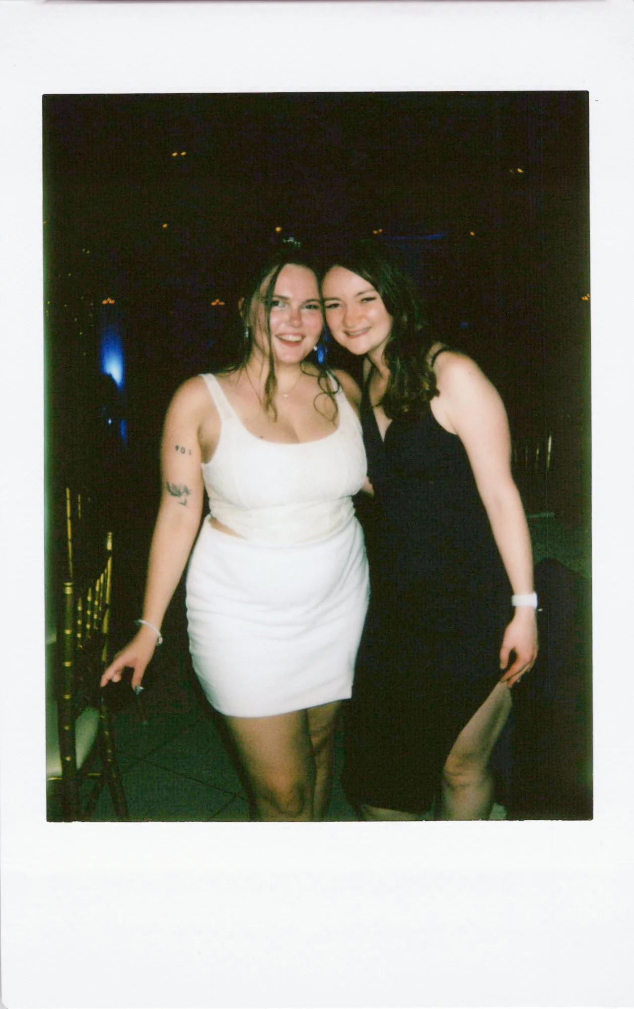 Two young women smiling and posing together at a nighttime event. One is wearing a white dress and the other a black dress. They are standing outdoors with a dark background and some lights visible.