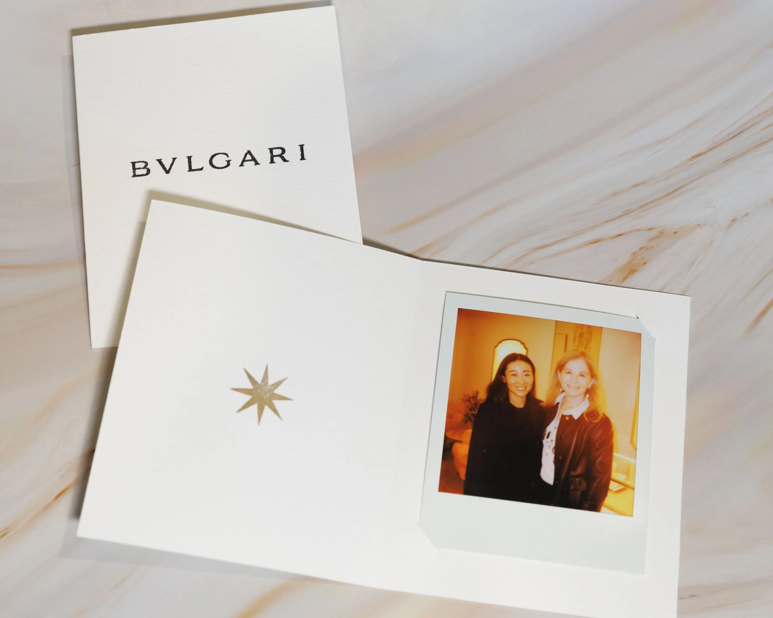 Close-up of a white card with the word "BVLGARI" printed on it, partially covering a photo of two women standing together, smiling, with a sunset background.