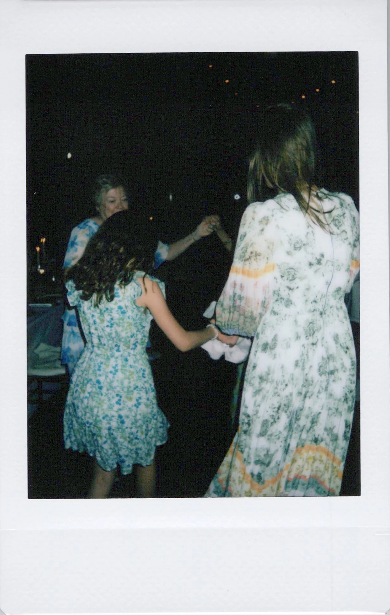 A woman in a white dress with a floral pattern is dancing with a girl in a blue and white dress. Another woman with blonde hair is dancing in the background. The setting appears to be at night with small lights visible in the dark background.