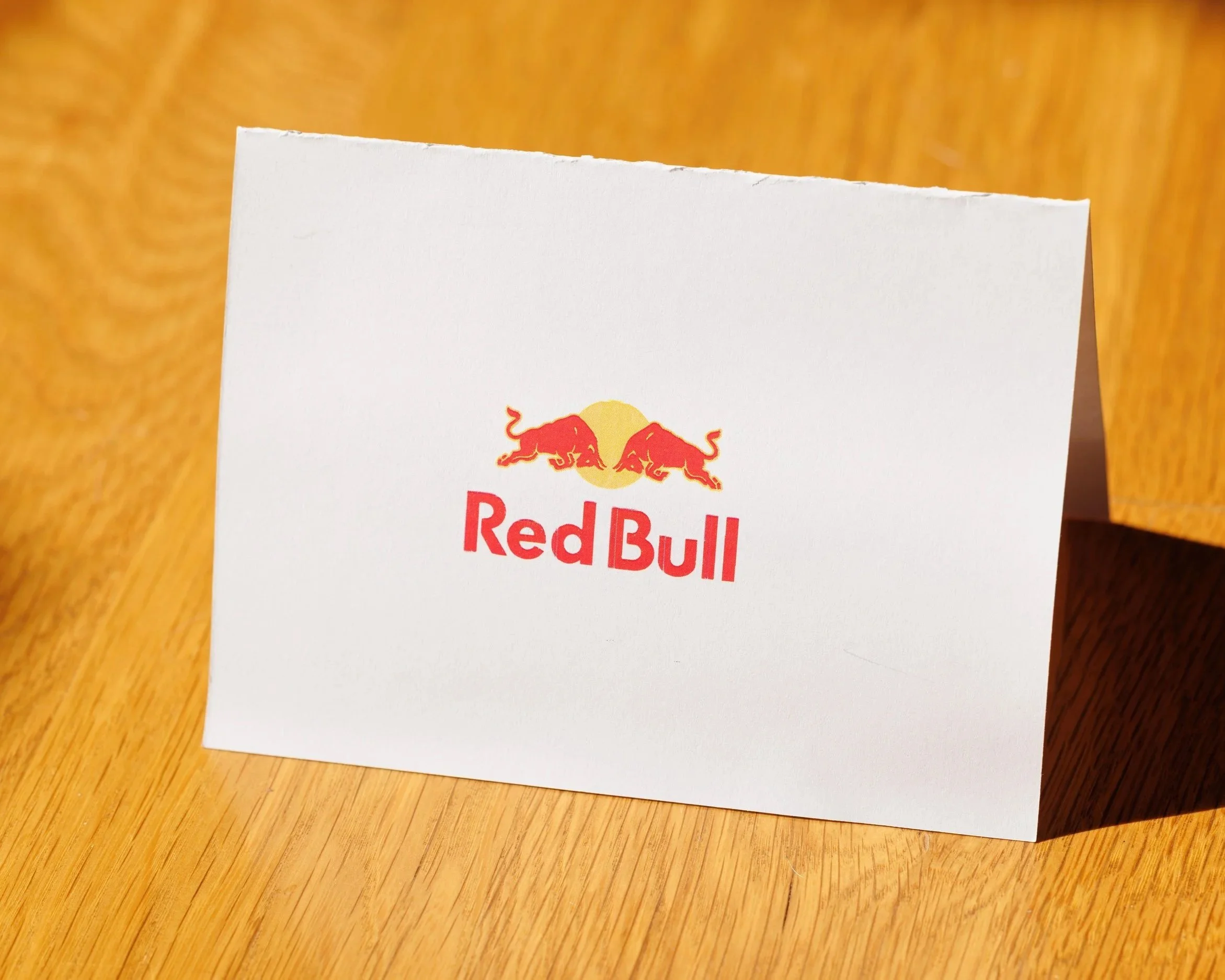 A white paper business card with the Red Bull logo featuring two red bulls fighting beneath a yellow sun, set on a wooden surface.
