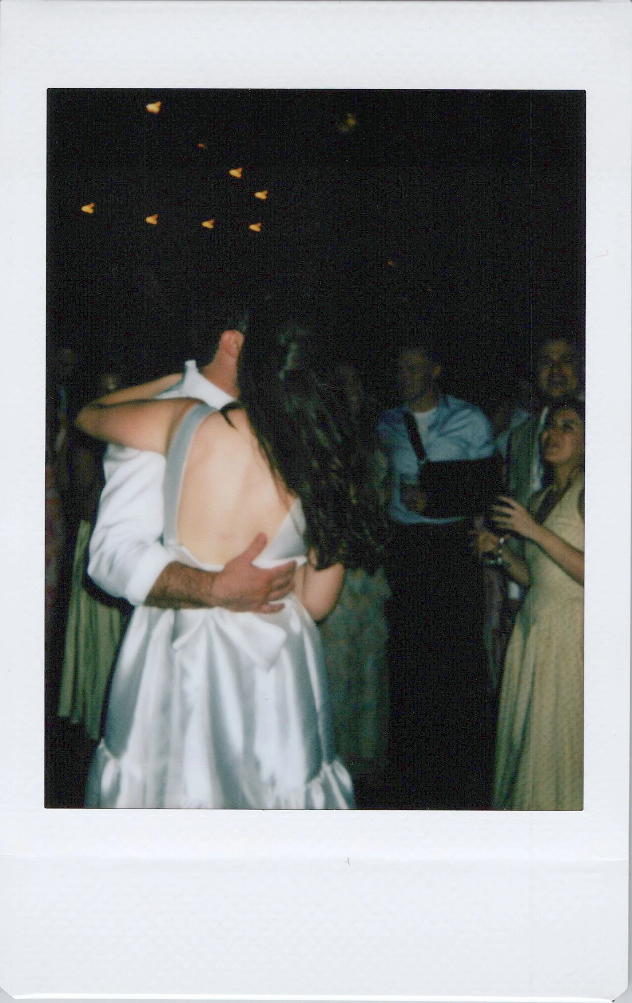 A newlywed couple dancing at their wedding reception with guests watching in the background. The bride is wearing a white wedding dress with an open back, and the groom is dressed in a white shirt.