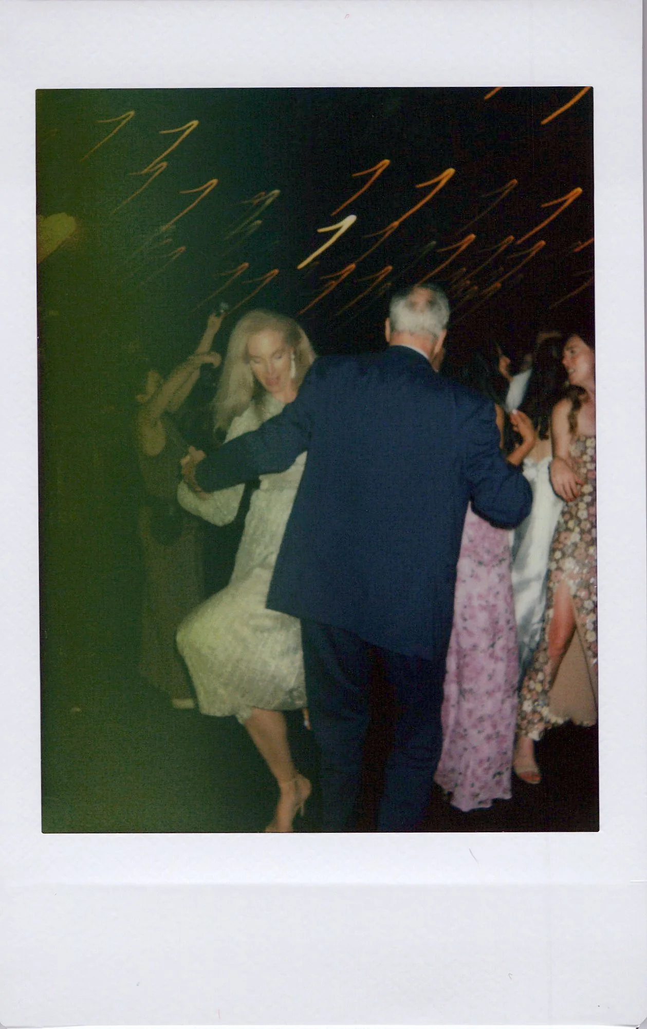 A woman in a light-colored dress dancing with a man in a dark suit at a party or celebration event. Several other women in formal dresses are seen in the background.